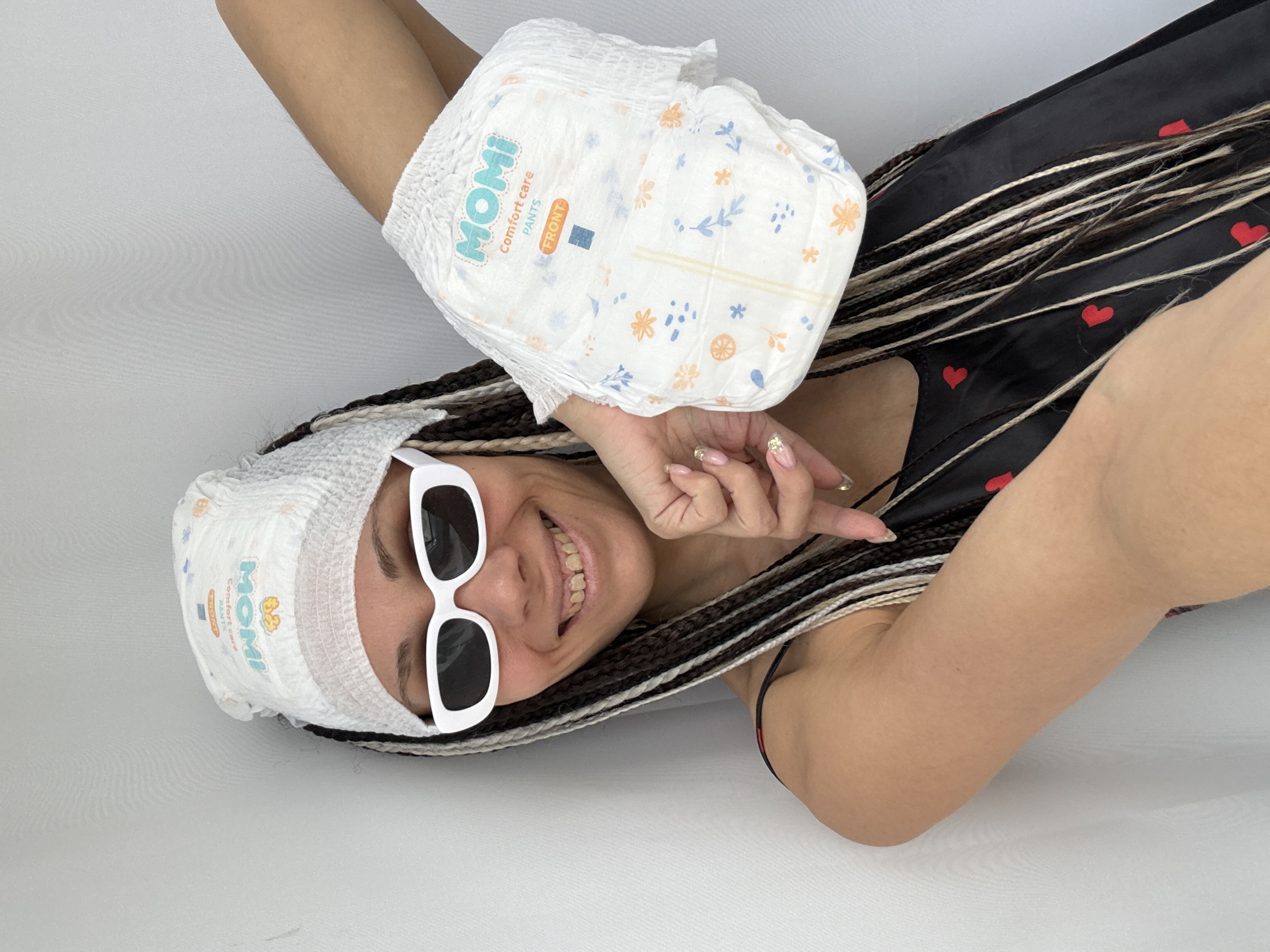 An overhead shot captures a smiling young woman posing against a plain white background. She is lying down, looking up at the camera with a wide, cheerful smile, revealing her teeth. She wears white, rectangular sunglasses perched on her nose and has long, multi-toned braided hair (dark, brown, and blonde/white strands).
One white baby diaper, adorned with light blue and orange floral and leaf patterns, is worn on her head like a hat. Another identical diaper is held up in her left hand, which is raised above her head. Her right arm is bent, and her hand, featuring manicured nails, is near her chin. She is wearing a dark garment, possibly a dress or top, which has small red heart patterns.
Visible text on the diapers includes "MOMI Comfort care PANTS" and "FRONT" with an arrow indicating direction. Smaller, less clear text like "TRUNK" or "TRONK" is also present, along with what appears to be a size indicator "L". The image exudes a playful and humorous mood. The setting appears to be indoors, possibly in a studio or a brightly lit room, with no specific outdoor or time-of-day cues. The context provided indicates the location is Rusanovo, Russia.