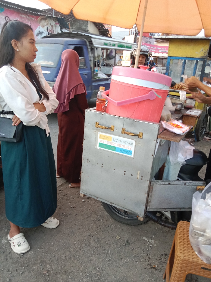 The image depicts a roadside scene in Sukomulyo, Indonesia, focused on a street food vendor. In the left foreground, a young woman with dark hair in a ponytail stands with her arms crossed, wearing a white long-sleeved blouse, a dark teal long skirt, and white Crocs. A black shoulder bag hangs from her left shoulder. She is looking towards the left, out of frame. Behind her, a person wearing a dark reddish-brown hijab and a matching long dress stands facing the vendor.
The central subject is a grey, metallic-sided food cart affixed to a motorcycle, indicated by a visible tire and engine part on the right. A large pink and white insulated cooler rests on the cart, partially obscured by an orange market umbrella extending upwards. To the left of the cooler, a clear plastic bottle containing an amber liquid is visible. A white sign affixed to the cart reads "BURJO KETAN HITAM WA: 088214879392 BERKAH." On the right side of the cart, a vendor's hands are actively handling small plastic bags and containers, appearing to prepare food or drinks. A person wearing a motorcycle helmet is partially visible behind the cooler and umbrella.
In the blurred background, a blue utility van or pickup truck is visible moving past, and other buildings and indistinct figures suggest a busy urban street. The ground is a mix of asphalt and dirt. In the extreme right foreground, a portion of a brown woven plastic stool or basket is visible, topped with clear plastic bags containing unidentified items.