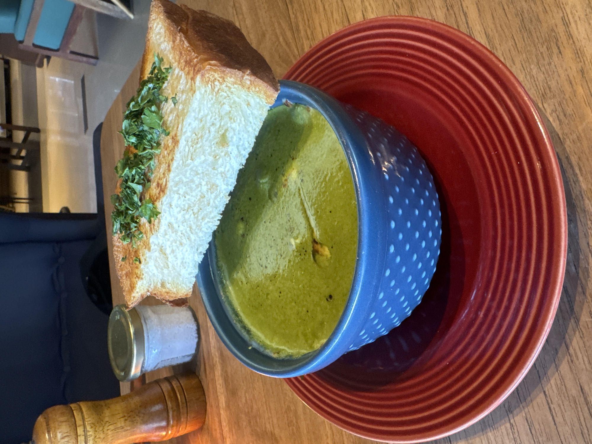 The image shows a bowl of green soup served with a piece of bread topped with green herbs. The soup is in a blue and white polka dot bowl which rests on a red plate with concentric rings. A small jar with a metallic lid and a wooden pepper grinder are situated next to the bread. The setting appears to be a restaurant or cafe with wooden tables. Based on the location, North Kuta, Indonesia, the food likely represents a dish commonly found in that region. No people are visible in the image.
