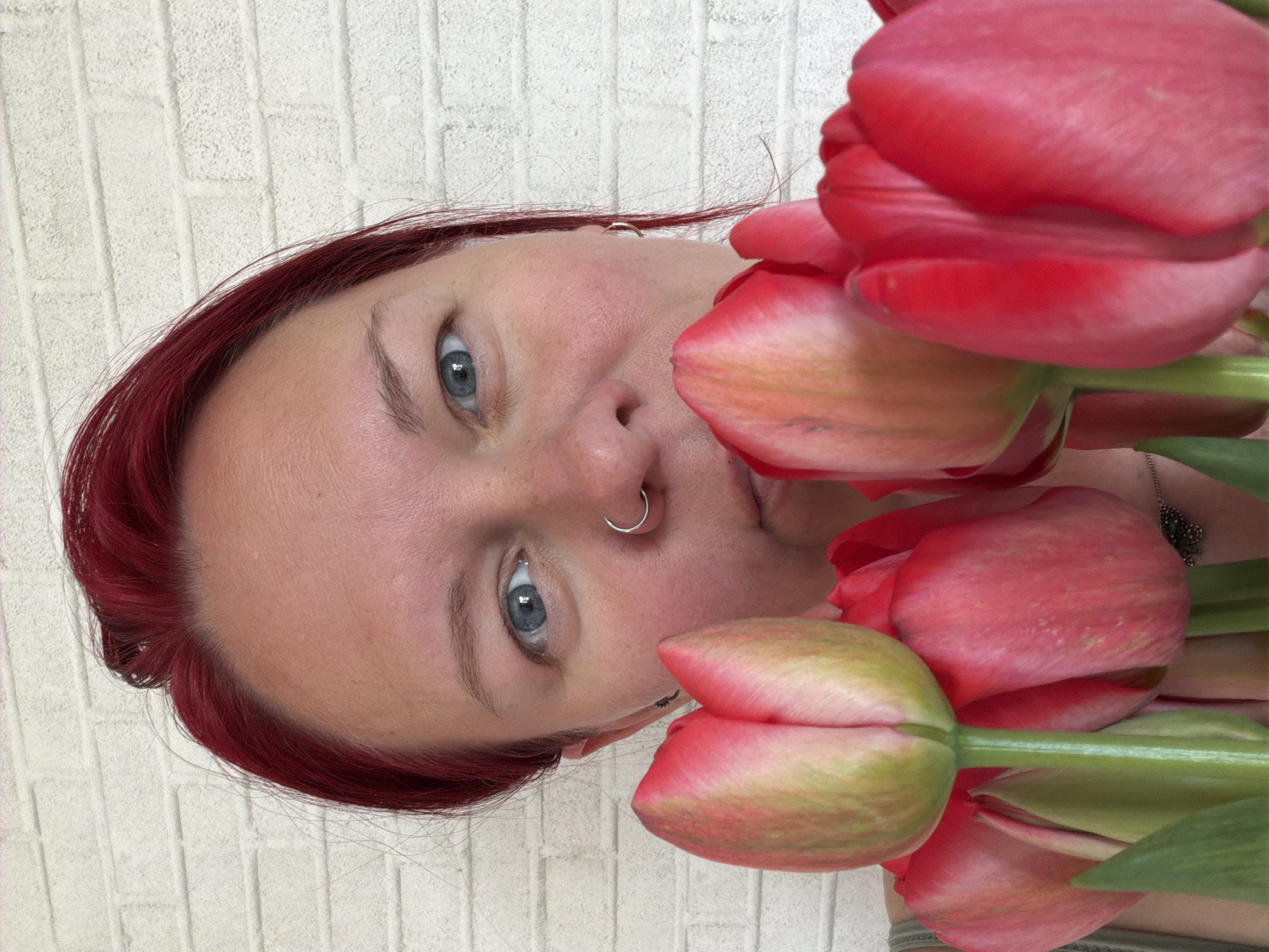 A woman with blue eyes and red hair is holding red tulips in front of a white brick wall. She has a nose ring. The image's location in Chaikovsky, Russia, is unknown.
