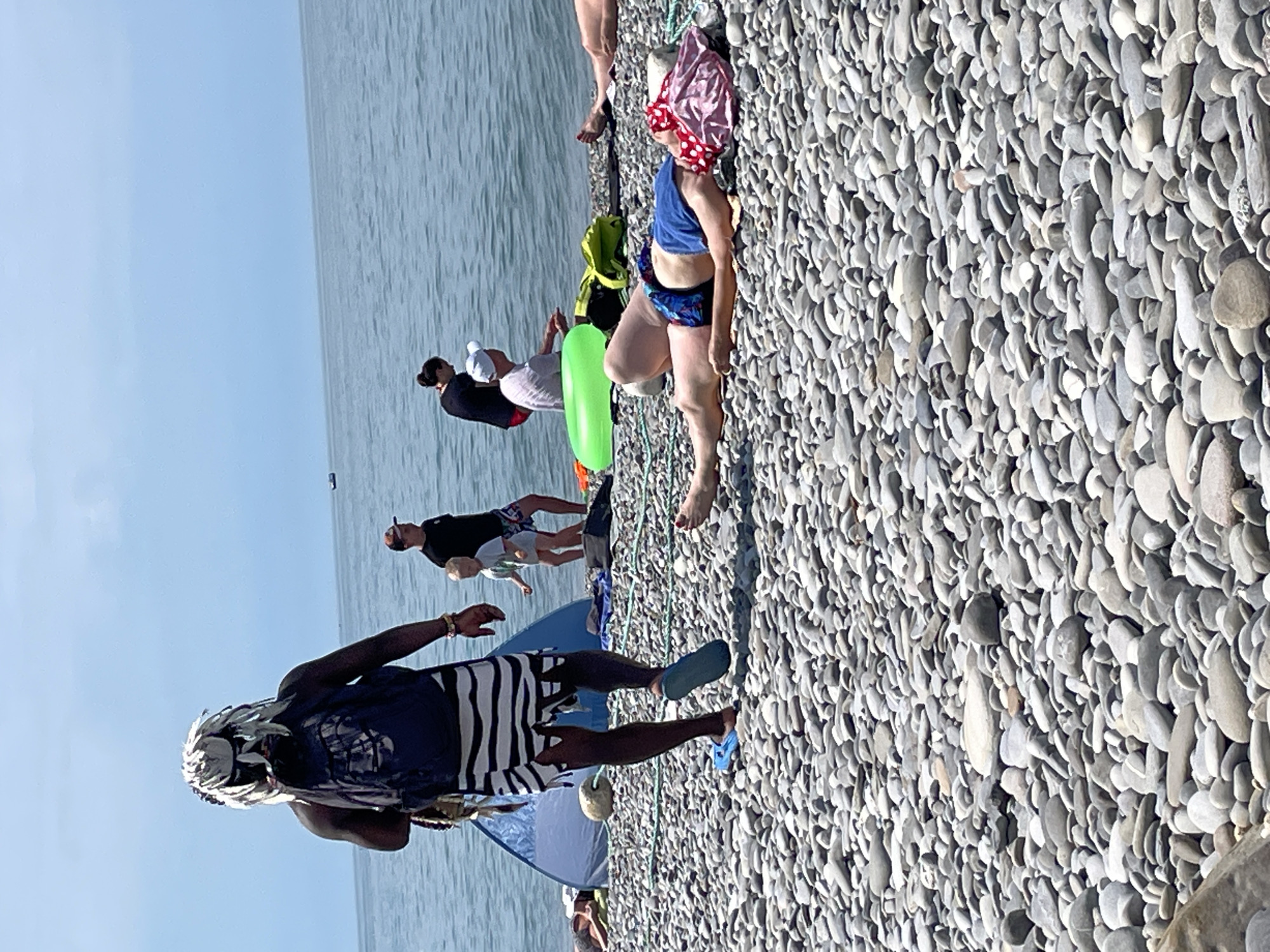 A group of people are on a pebble beach by the sea. In the foreground, a person wearing a headdress with feathers, a backpack, and striped shorts walks away from the camera. They are wearing blue flip-flops. To the right, a person is lying on their back on a towel, wearing a blue top and patterned shorts. A pink and red fabric is on their head. Behind them, a green inflatable ring is on the ground. Further back, a woman in a black top and red shorts is standing near the water. Beside her, a man in a white t-shirt and a white cap is sitting down. A young child in a grey t-shirt is standing between them. A blue tent is visible behind the child. The sea is calm and a light blue sky is above.