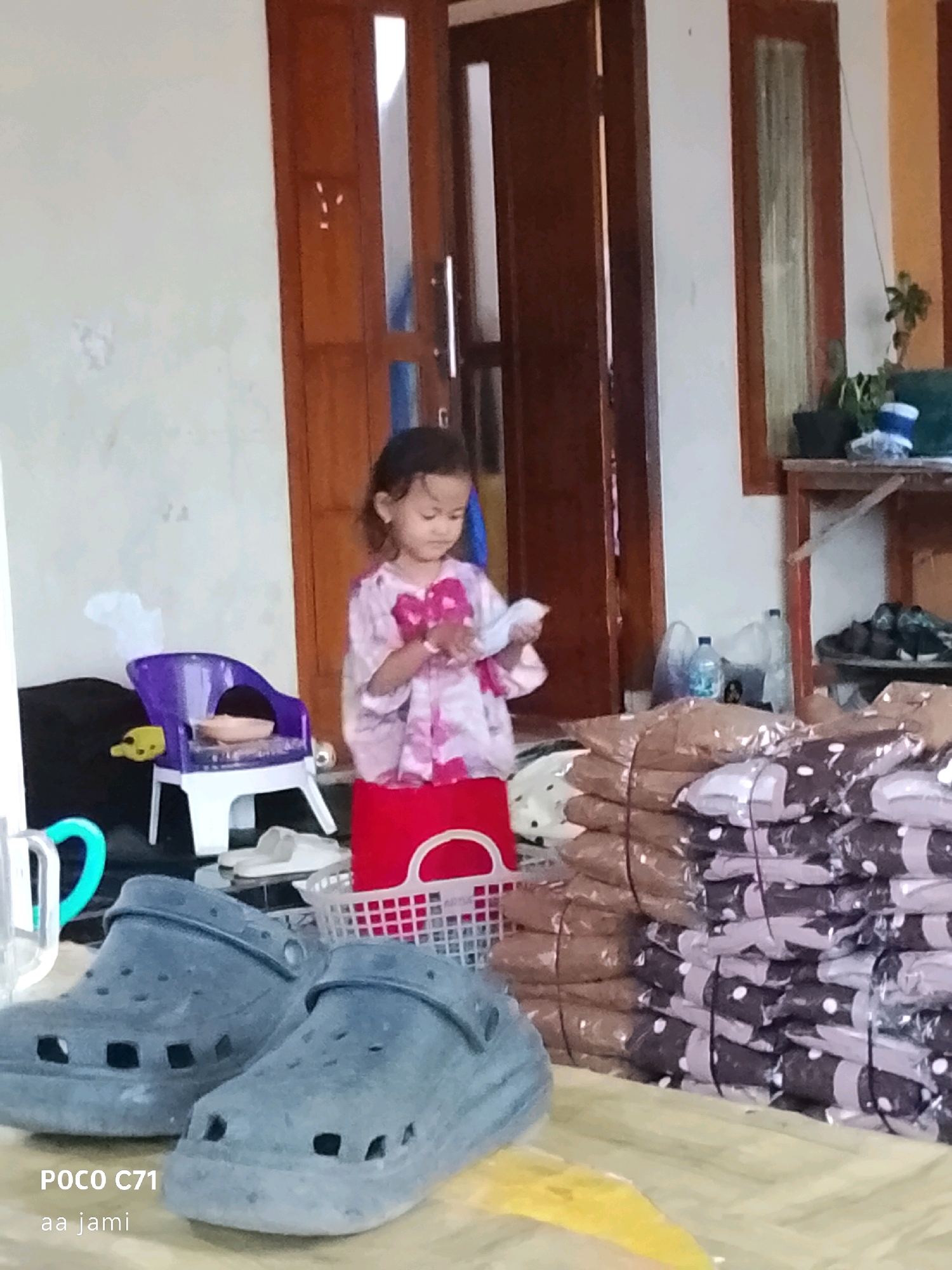 A young girl wearing a pink and white patterned dress is standing in front of a red basket. She is holding a white object in her hands. To her left is a small purple chair with a white base. In the foreground, there are two large gray clogs. Behind the girl, stacks of goods wrapped in plastic are visible, some with a dark pattern and white polka dots. The scene appears to be outdoors, possibly in a residential or commercial area, with a wooden door in the background. The image has a watermark indicating it was taken with a POCO C71 phone and includes the text "aa jami". This description is likely of a scene in Tasikmalaya, Indonesia, given the provided location information.