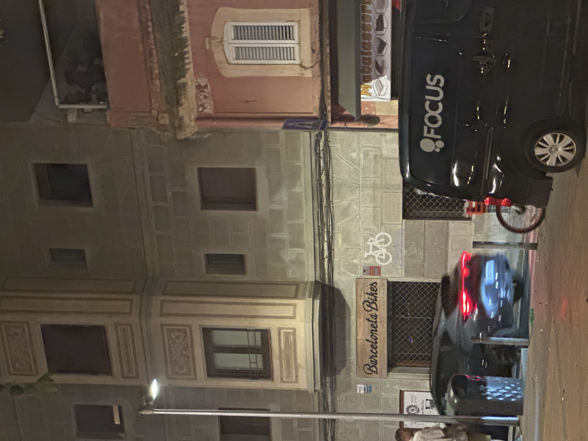 The image depicts a street scene in Barcelona, Spain, at night. A black van with the logo "FOCUS" is parked on the right side of the frame. To the left of the van, a black car with its brake lights illuminated is also parked. A bicycle is visible in the foreground on the right. In the background, a building features a sign that reads "Barceloneta Bikes" with a bicycle graphic next to it. Another building with a pink facade and a white-shuttered window is visible to the right of the "Barceloneta Bikes" building. A street lamp illuminates the scene from the left side of the frame. A trash can is positioned near the parked car.