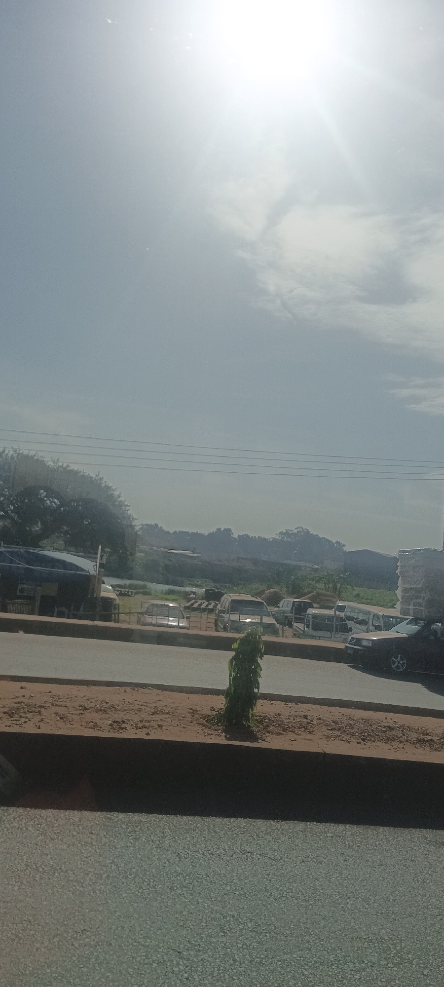The image shows a street scene in Bukuru, Nigeria, on a bright day. The foreground features a gray paved road and a low, reddish-brown strip of earth planted with a small, leafy green shrub. Beyond this strip is a busy street with several vehicles parked and possibly moving. The vehicles include vans, cars, and other utility vehicles. In the background are various structures, possibly commercial or industrial, including some covered by a dark tarp or roof. The distant background consists of hilly or elevated terrain covered with vegetation. Power lines are visible stretching across the upper part of the image. The sun is high and bright, partially obscured by a thin layer of clouds, causing a strong glare.
