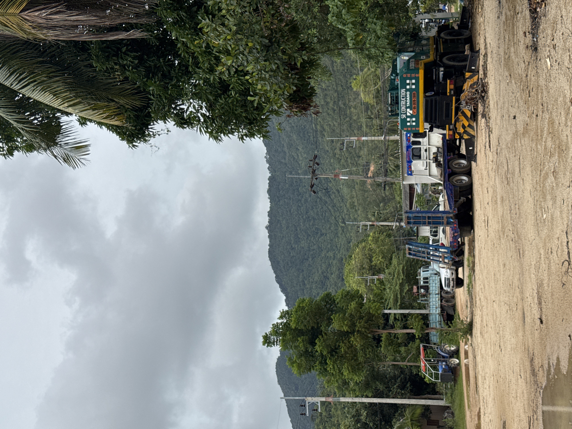 The image depicts an outdoor scene in a lot near the coast of Koh Phangan, Thailand, under a cloudy sky. Several vehicles are parked on a sandy, uneven surface, with some puddles of water indicating recent rain. In the foreground, a large green crane truck with "SR CONSTRUCTION TADANO" branding is prominent, its outriggers extended. To its left, a flatbed trailer is attached to a white truck. Further left, a blue flatbed truck is parked, and a small blue tractor with a red frame is visible near the edge of the lot. In the background, lush green hills dominate the landscape, with scattered trees and utility poles in between. The overall impression is one of a construction or transportation yard.