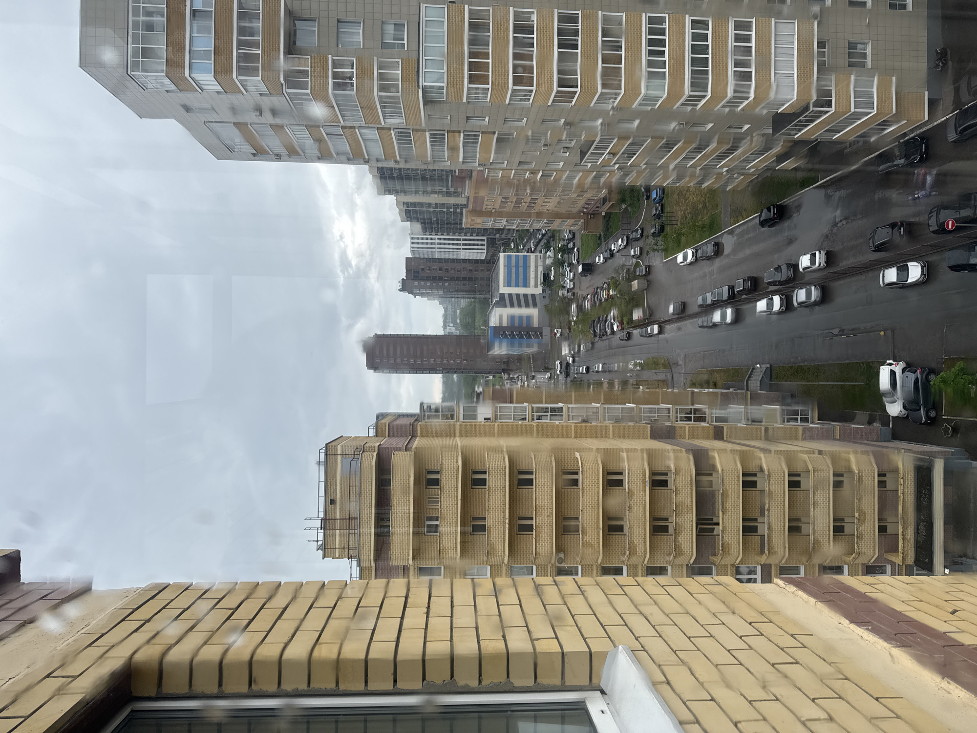 Here is a detailed description of the image:
**Environment:** The image captures a street scene on a rainy day, as evidenced by the wet pavement and overcast sky. The perspective is from an elevated position, likely from a window within a high-rise building.
**Location:** The location appears to be an urban area, likely a residential district with multi-story apartment buildings lining both sides of a road.
**Buildings:** Several high-rise apartment buildings are visible. The buildings feature brick and concrete facades.
**Road:** The road is filled with cars, indicating moderate traffic flow. The vehicles are various colors, with white and dark-colored cars.
**Objects:**
* Cars parked and driving on the wet road.
* Brick buildings.
* Windows.
* A window from the location where the image was taken.
**People:** There is no specific indication of pedestrians or individuals visible in this image, as the focus is on the urban landscape and traffic.
**Actions:** Cars are moving along the wet road.
**Weather:** Rainy.