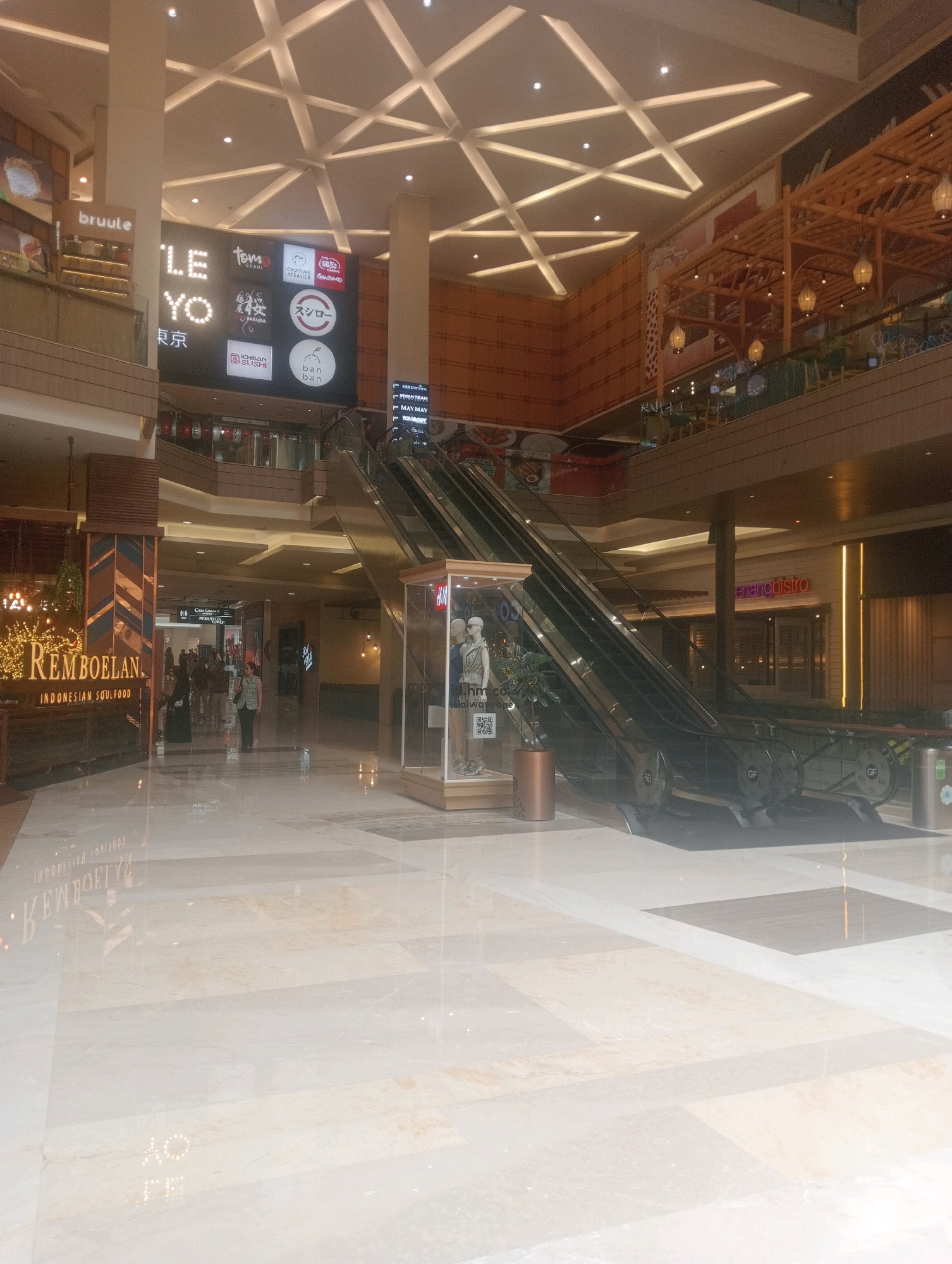 The image captures a wide-angle view of a modern, multi-level shopping mall interior. The scene is well-lit, with a combination of ambient ceiling lights and spotlights illuminating the space. In the foreground, a polished marble floor reflects the overhead lighting and architectural elements.
Dominating the center of the frame are escalators leading to an upper level. To the left of the escalators, a sign with the text "REMBOELAN INDONESIAN SOULFOOD" is prominently displayed, suggesting a restaurant or food court area. Further left, a sign with the word "bruule" is visible.
Above the escalators, a large digital screen displays various logos and text, including "LE YO TOKYO," "TOMO SUSHI," and others, indicating a collection of food and beverage establishments. Above this digital display, a complex, geometric ceiling structure made of white beams is visible.
To the right of the escalators, a store with a large glass display window features mannequins dressed in casual clothing, identified by a partial "H&M" logo. Further to the right, a sign with "penang bistro" is visible.
In the middle ground, several people are walking or standing, blurred due to the image quality or motion. The overall environment suggests a bustling commercial space with a focus on dining and retail. The architectural style is contemporary, with clean lines and a neutral color palette accented by wooden elements and signage. The context provided places this scene in a shopping mall in South Jakarta, Indonesia.