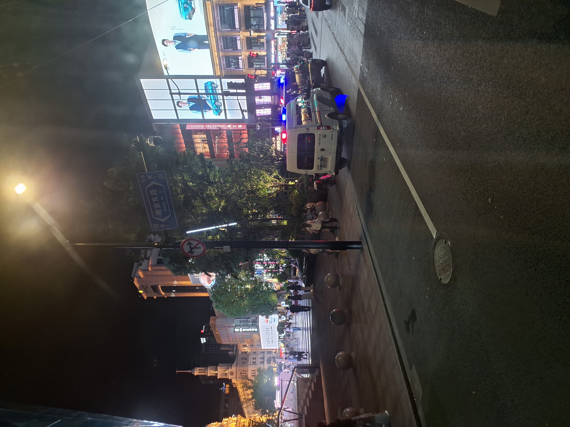 The image depicts a nighttime street scene in Shanghai, China, specifically on Nanjing Road East, as indicated by a street sign. The scene is illuminated by a mix of streetlights, building lights, and large digital billboards. In the foreground, a paved road with a white line marking is visible, with a manhole cover on the right. To the left of the road, a pedestrian area with decorative spherical bollards and a tiled surface is present. Several people are walking in this area, and some are seated on benches.
Dominating the middle ground are two law enforcement vehicles, a white van and a dark SUV, both equipped with flashing red and blue lights, suggesting an ongoing police presence or activity. The white van has Chinese license plates and appears to be parked. Behind the vehicles, a large building with multiple floors is visible, featuring illuminated windows and commercial signage. A prominent digital billboard displays images of two men in suits and a blue car. Trees and other greenery are scattered throughout the scene. Further in the background, more buildings and pedestrian activity are observable, contributing to a busy urban environment. The overall atmosphere is that of a well-lit, active city street at night.