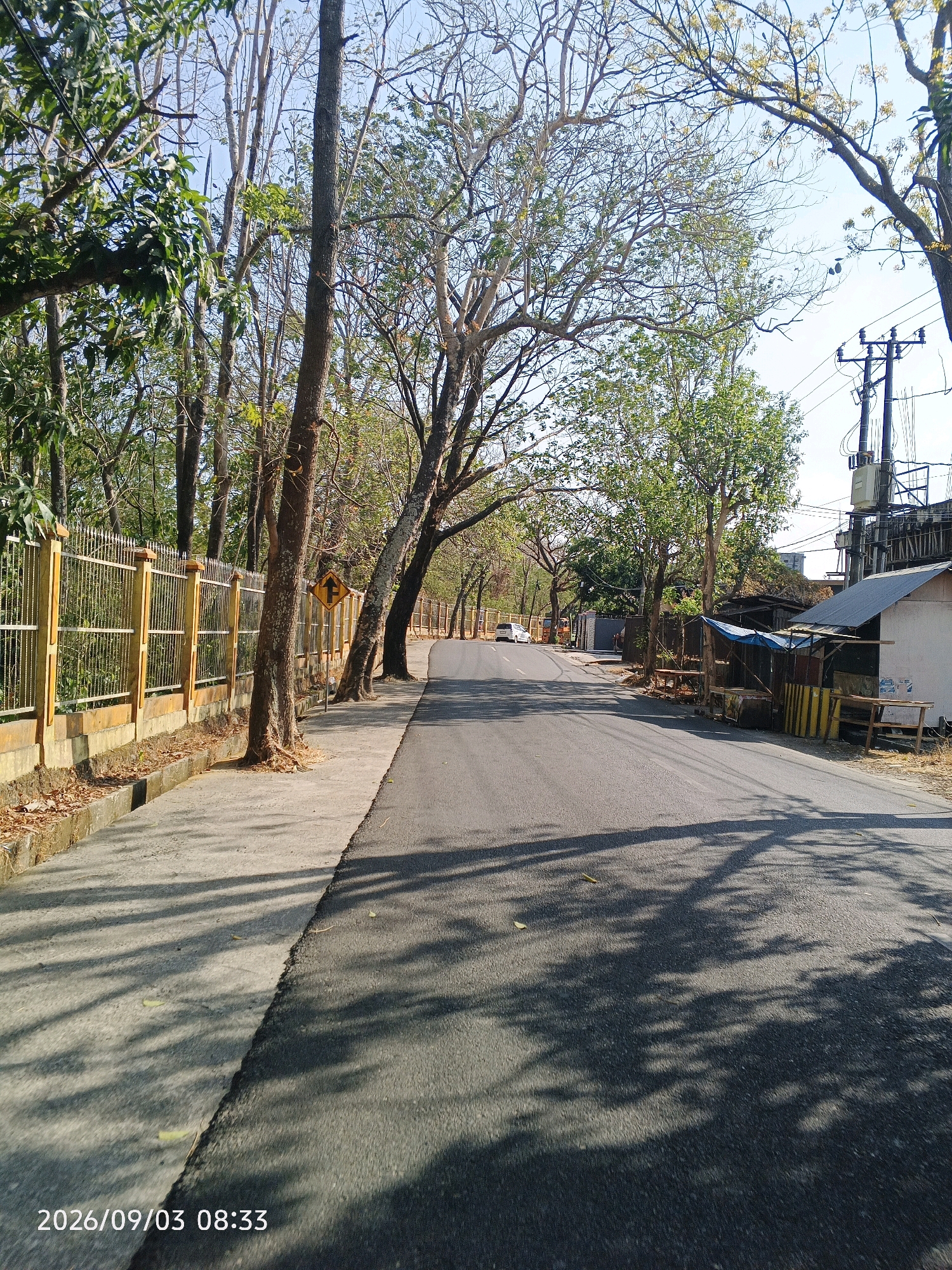 The image shows a paved road lined with trees and a sidewalk. On the left side of the road, there is a yellow metal fence and a sidewalk with scattered leaves. The road continues into the distance, curving slightly to the right. On the right side of the road, there are a few small structures with corrugated metal roofs and tarps, and some wooden stalls. A utility pole with wires is also visible. The sky is clear and blue. The timestamp "2026/09/03 08:33" is visible in the lower-left corner. The scene appears to be a roadside in a semi-urban or suburban area, likely with sparse vegetation. The image does not provide enough information to pinpoint a specific location within Pare-Pare, Indonesia.