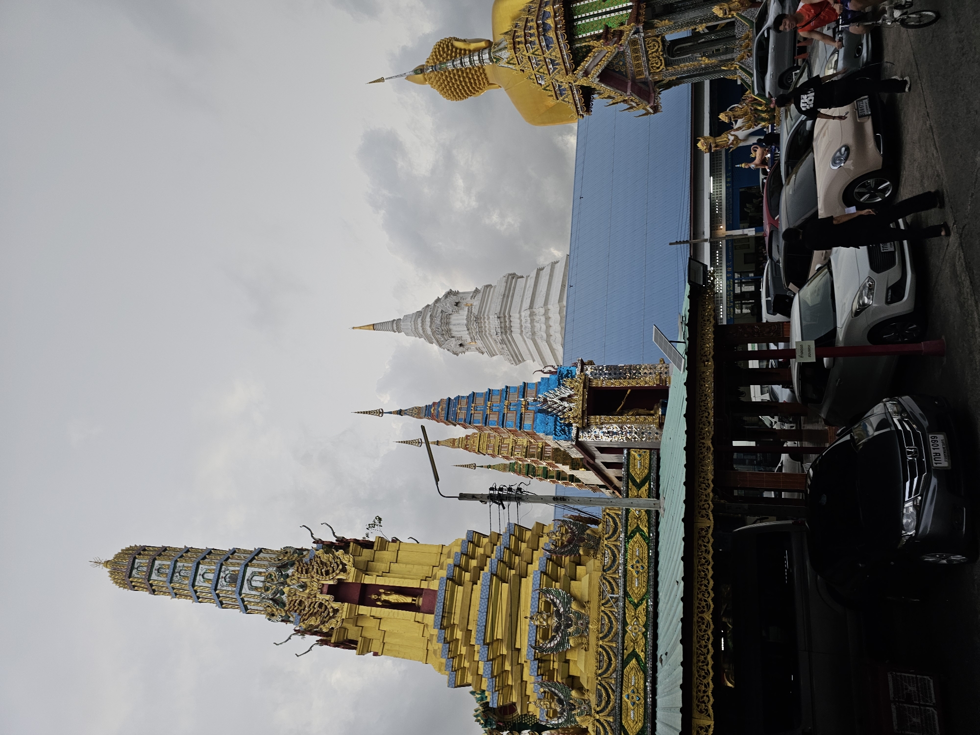 The image depicts a scene in front of a Buddhist temple complex in Bangkok, Thailand, under an overcast sky. Dominating the left side of the frame is a tall, ornate golden stupa, adorned with intricate carvings and multiple tiers. To the right of this, several smaller, colorful stupas in blue, gold, and red also stand prominently. Further to the right, a colossal golden statue of Buddha's head and upper torso is visible, facing away from the camera. In the foreground, several cars are parked, including a dark sedan with a license plate displaying "108 1099". Three individuals are present: one person on a bicycle on the far right, another standing near the parked cars, and a third person walking away from the viewer, dressed in black. A fourth individual, also in black, is standing closer to the cars. The background features a building with a blue roof, and various architectural elements typical of Thai temples. The overall atmosphere is somber due to the cloudy weather, but the vibrant colors of the temple structures provide a stark contrast.