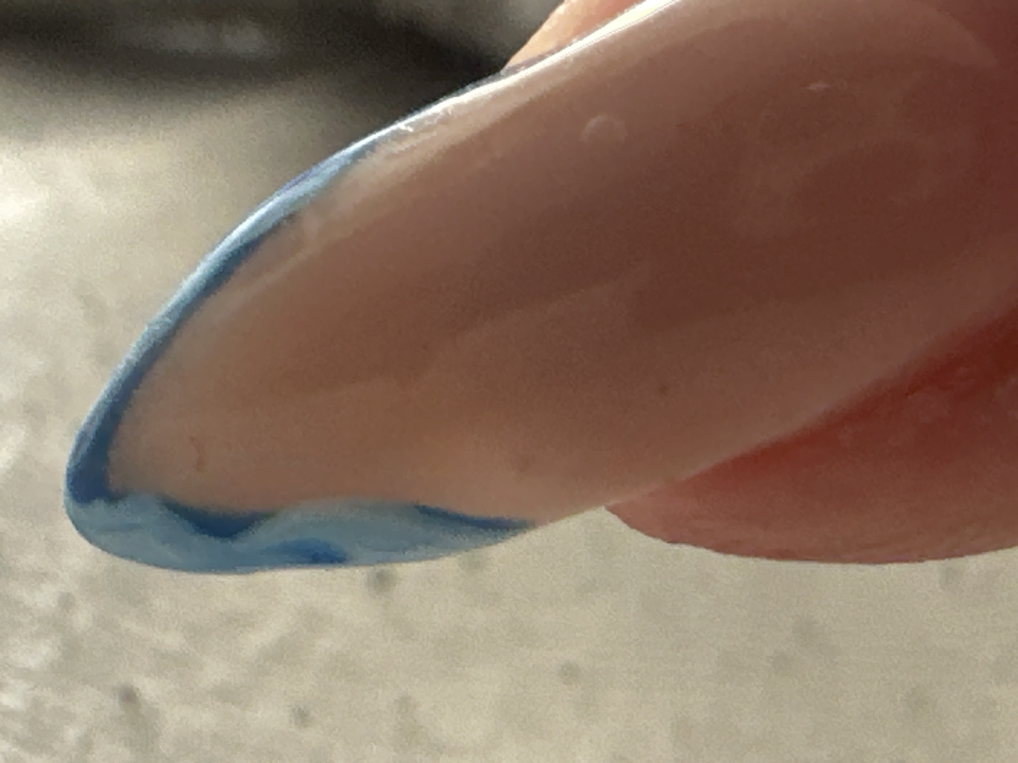 The image shows a close-up view of a fingernail with blue and white nail polish applied to the tip, creating a marbled or wave-like effect. The nail is attached to a finger with light-toned skin. The background is out of focus and appears to be a neutral, textured surface. There is no indication of a location in Shumen, Bulgaria, or any specific activity occurring.