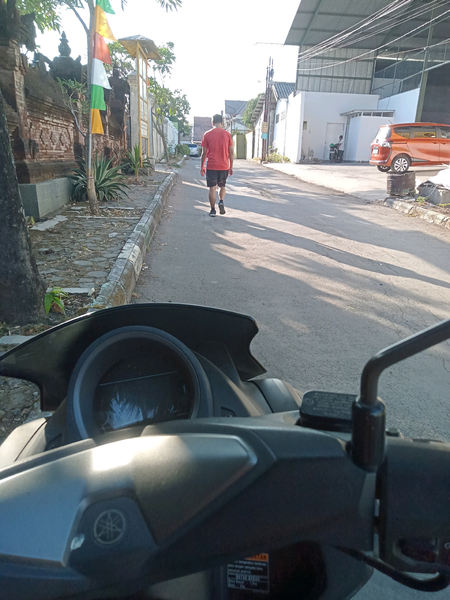 The image depicts a street scene in Cirebon, Indonesia, viewed from the perspective of a motorcycle. In the foreground, the handlebars, instrument panel, and part of the motorcycle's fairing are visible. A Yamaha logo is present on the fairing. The street is paved with asphalt and has a curb on the left side. A person in a red t-shirt and black shorts is walking away from the camera down the street. The left side of the street features a stone-paved sidewalk, a traditional brick wall with ornate carvings, and lush green plants. Several colorful flags, white, green, and orange, are attached to a pole near the wall. Further down the street, buildings with white walls and corrugated metal roofs are visible. An orange car is parked on the right side of the street. The lighting suggests it is either early morning or late afternoon, with long shadows cast across the road.