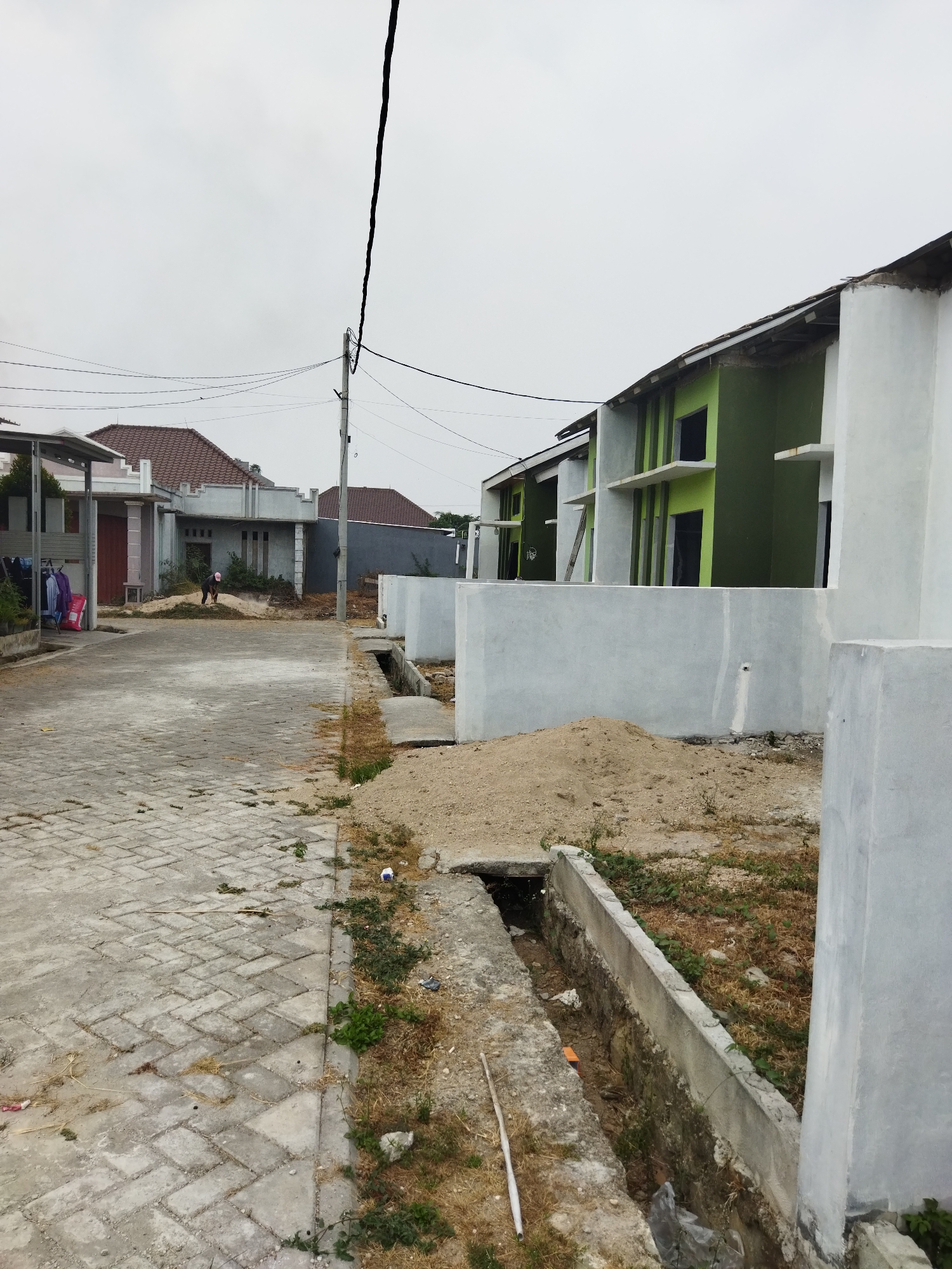 In Suka Sari, Indonesia, an image captures a view down a paved walkway bordered by construction sites. The walkway, made of interlocking grey bricks, shows signs of wear and is partially covered in dry grass and debris. To the left, a low building with a red-tiled roof and white columns stands, with laundry hanging on a rack outside. Further down the walkway, a person wearing a pink head covering is bent over, appearing to be tending to the grass.
On the right side of the walkway, construction is evident with several newly built houses in various stages of completion. These houses feature green and white exteriors, with visible window openings and concrete walls. A pile of sand and a narrow concrete drainage ditch run along the front of these properties. A utility pole with electrical wires is also present, extending upwards into a cloudy sky. The environment appears to be a residential development undergoing construction.