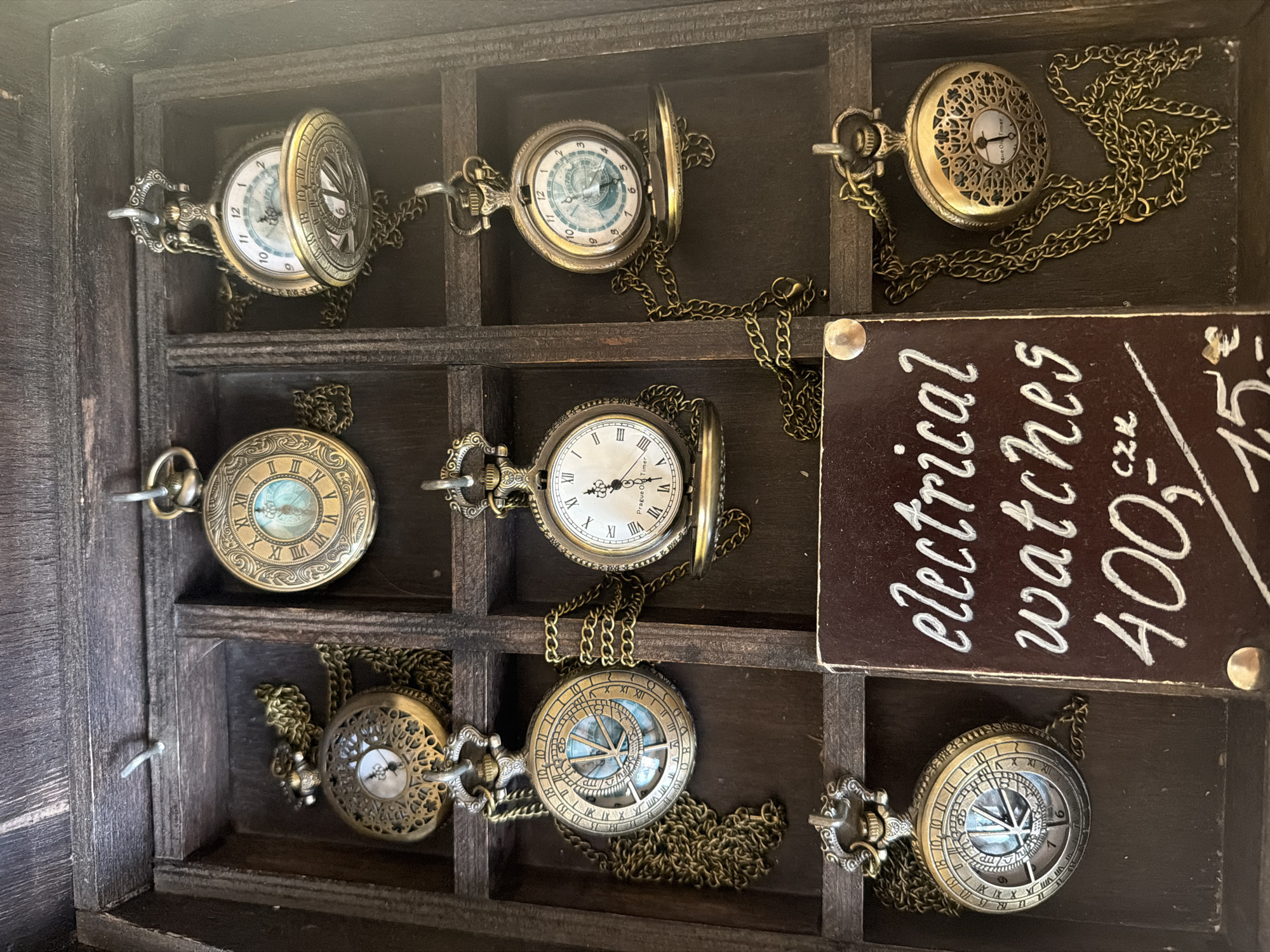 In a wooden display case with nine compartments, a collection of eight antique pocket watches with chains are arranged. Some watches are closed, while others are open, revealing their faces. The case is situated in a shop in Prague, Czechia, where a sign with white lettering on a dark brown background reads "electrical watches 400 czk 15€". The overall scene evokes a sense of vintage charm and historical interest.