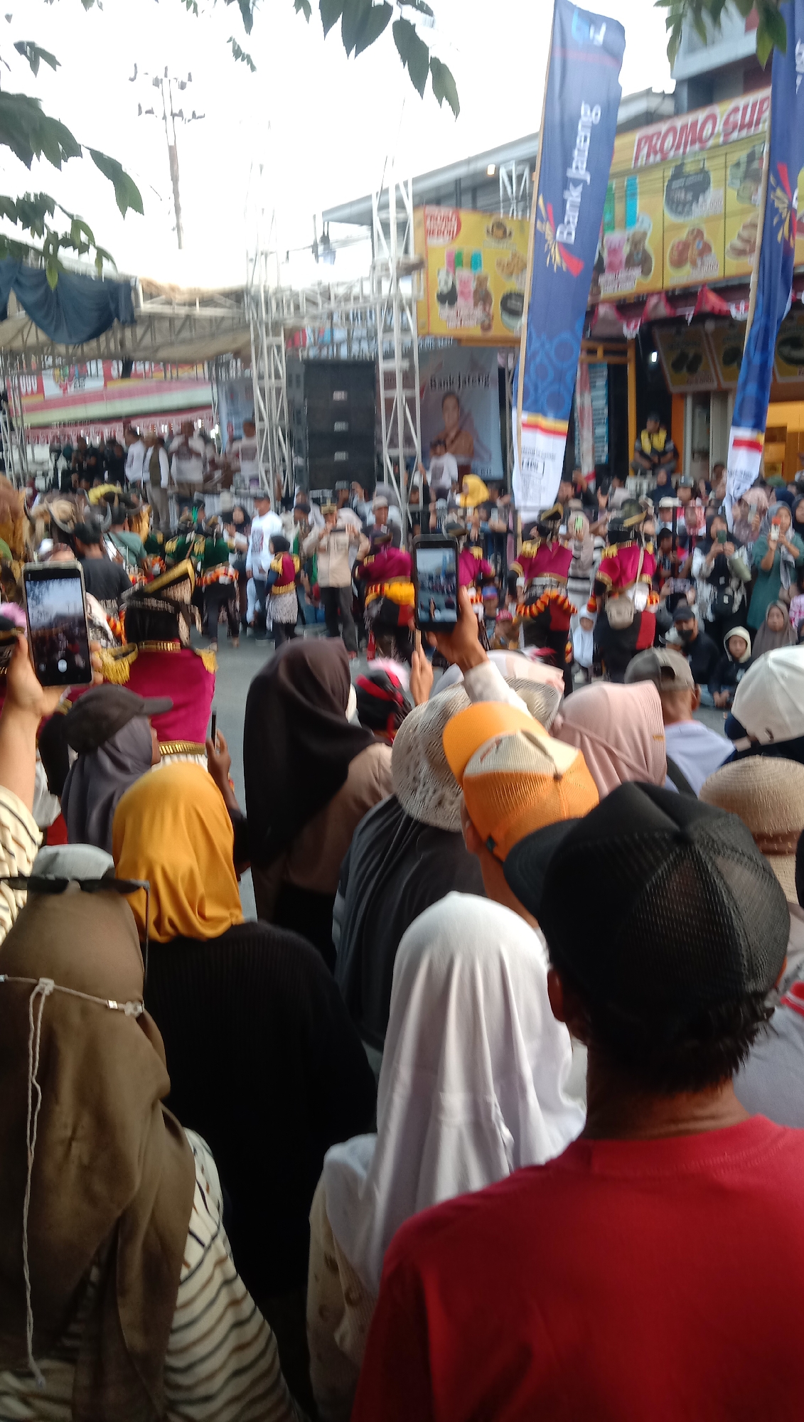 This image captures a lively street scene in Jatisawit, Indonesia, likely during a cultural event or parade. A large crowd of onlookers is gathered, many holding up smartphones to capture the spectacle. In the background, a stage or performance area is visible, complete with scaffolding, speakers, and banners. One prominent banner reads "Bank Jateng," suggesting a local bank's sponsorship. Several individuals in traditional or ceremonial attire, possibly performers, are visible in the distance, adding to the festive atmosphere. The environment appears to be an outdoor urban setting, with trees and overhead wires suggesting a street or public square. The overall impression is one of community engagement and celebration.