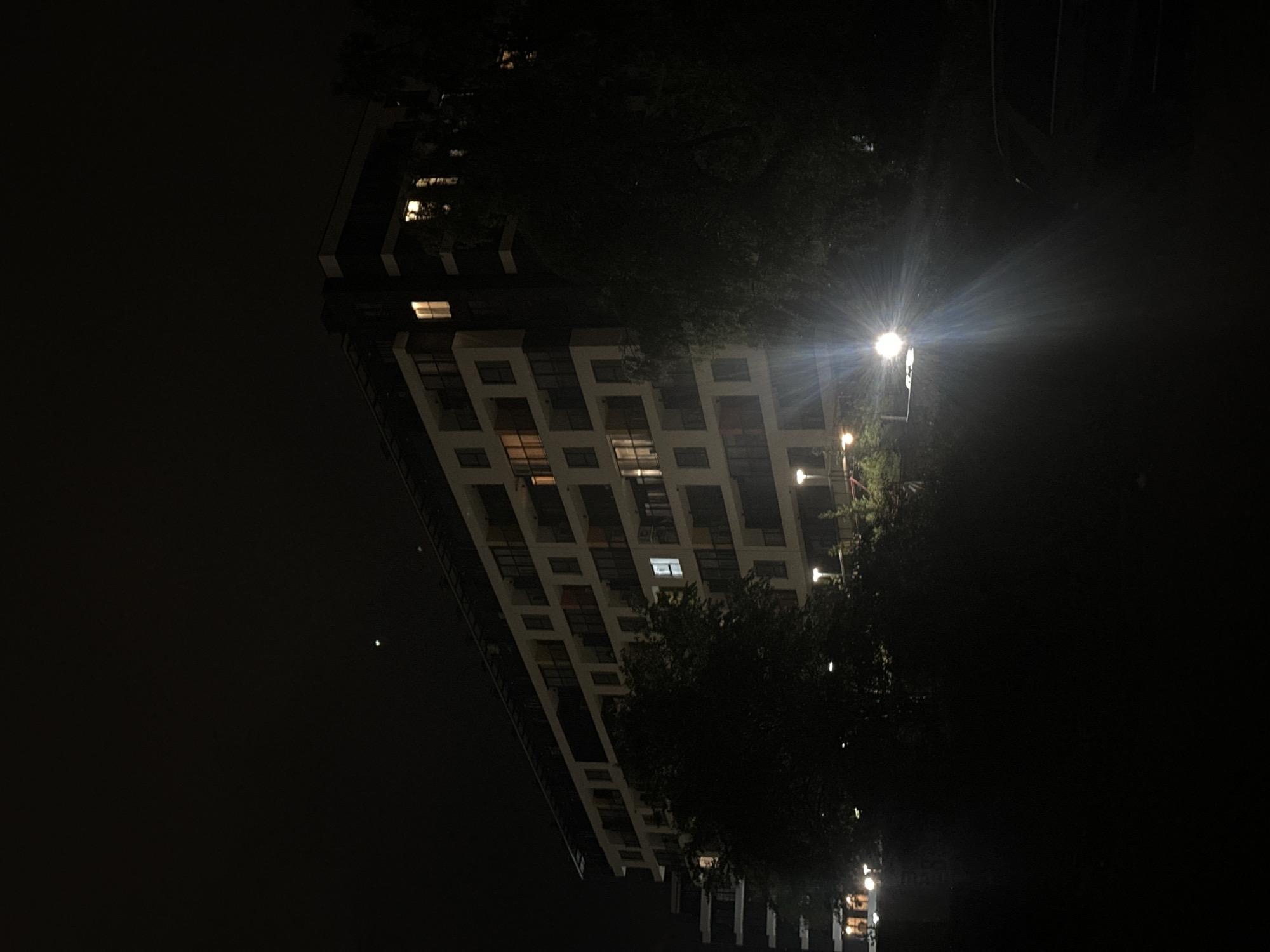 The image shows a tall building at night. The building has a grid-like facade with illuminated windows scattered across its surface. A street light with a strong glare is located to the right of the building, partially obscured by trees. The surrounding environment is dark, with the building and street lights providing the main sources of illumination. The exact location within Sofia, Bulgaria cannot be determined from this image.