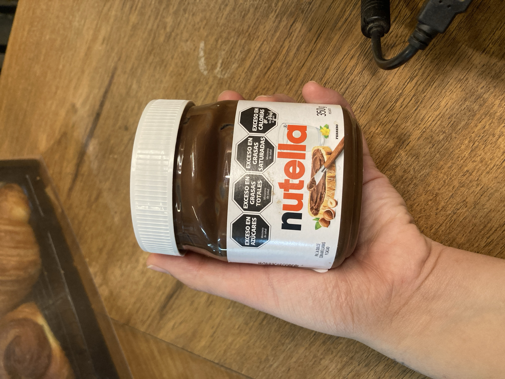 A close-up, vertical shot shows a person's hand holding a jar of Nutella spread, positioned diagonally across a wooden surface. The hand grasps the jar, which is mostly full of the dark brown hazelnut spread. The jar features a white ribbed lid. On the label, the prominent "nutella" logo is visible, along with "FERRERO" and "350g PESO NETO" indicating its weight.
The label also displays several hexagonal warning seals, common in some countries, indicating high levels of certain ingredients:
* "EXCESO EN AZÚCARES" (Excess in Sugars)
* "EXCESO EN GRASAS TOTALES" (Excess in Total Fats)
* "EXCESO EN GRASAS SATURADAS" (Excess in Saturated Fats)
* "EXCESO EN CALORIAS" (Excess in Calories)
Each warning is accompanied by "Ministerio de salud" (Ministry of Health). Below the main logo, text reads "PASTA DULCE CON AVELLANAS Y CACAO" (Sweet spread with hazelnuts and cocoa).
In the background, a dark wooden tabletop with visible grain is seen. To the upper right, a black USB cable is partially visible. On the left, a corner of a clear plastic container, possibly holding pastries or a croissant, can be glimpsed. The lighting is even, suggesting an indoor setting.