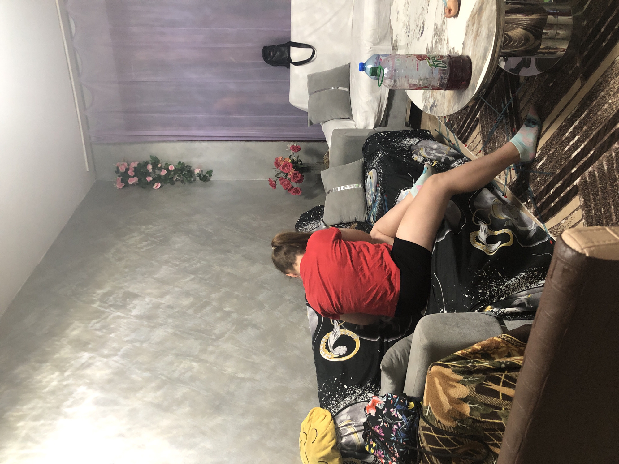 In an indoor living room setting, a young person with light brown hair pulled into a ponytail is seated on a couch. They are wearing a red t-shirt and black shorts, with light blue and pink socks. The couch is adorned with a black and white patterned throw blanket. To the left of the person, a yellow smiley face pillow and a floral patterned bag are visible. The wall behind the couch features a textured, light gray finish. Adjacent to this couch is a light-colored couch with gray pillows. In the background, sheer purple curtains hang across a window. On a coffee table in front of the couches, a large plastic bottle containing a dark red liquid and a second smaller blue-capped bottle are placed. A patterned rug covers the floor. A floral arrangement in a vase is visible on a surface to the left of the white couch.