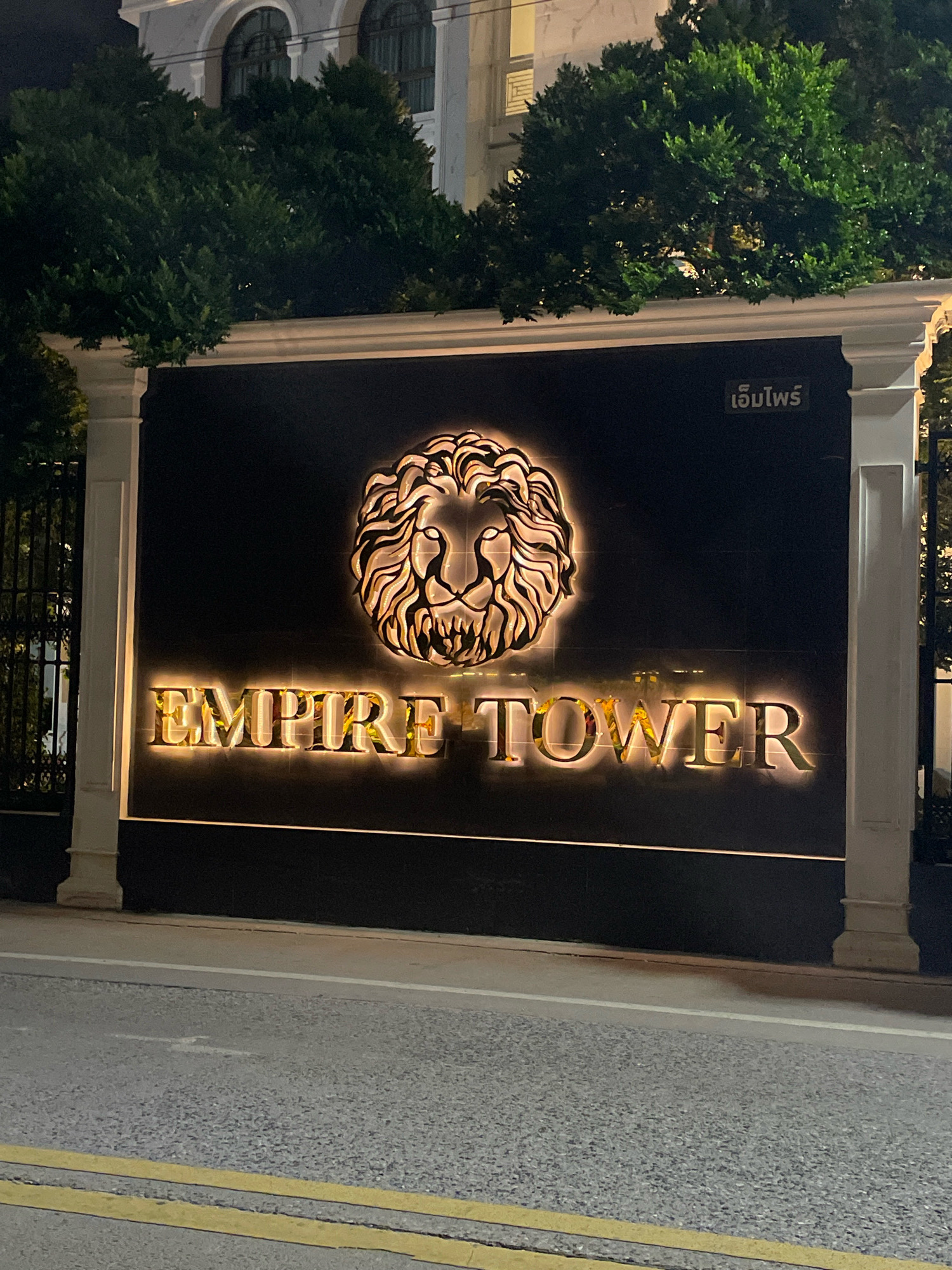 The image shows a black sign with illuminated text and a logo. The text reads "EMPIRE TOWER" in large, gold-colored, illuminated letters. Above the text is a stylized logo of a lion's head, also illuminated with a warm, golden light. To the right of the lion logo, there is smaller text in a light color that appears to be in Thai script. The sign is framed by two white pillars. In the background, there are lush green trees and a building with arched windows. The sign is situated outdoors, with a street visible in the foreground, indicated by asphalt and yellow road markings. The lighting suggests it is nighttime. This is the entrance sign for Empire Tower in Pattaya City, Thailand.