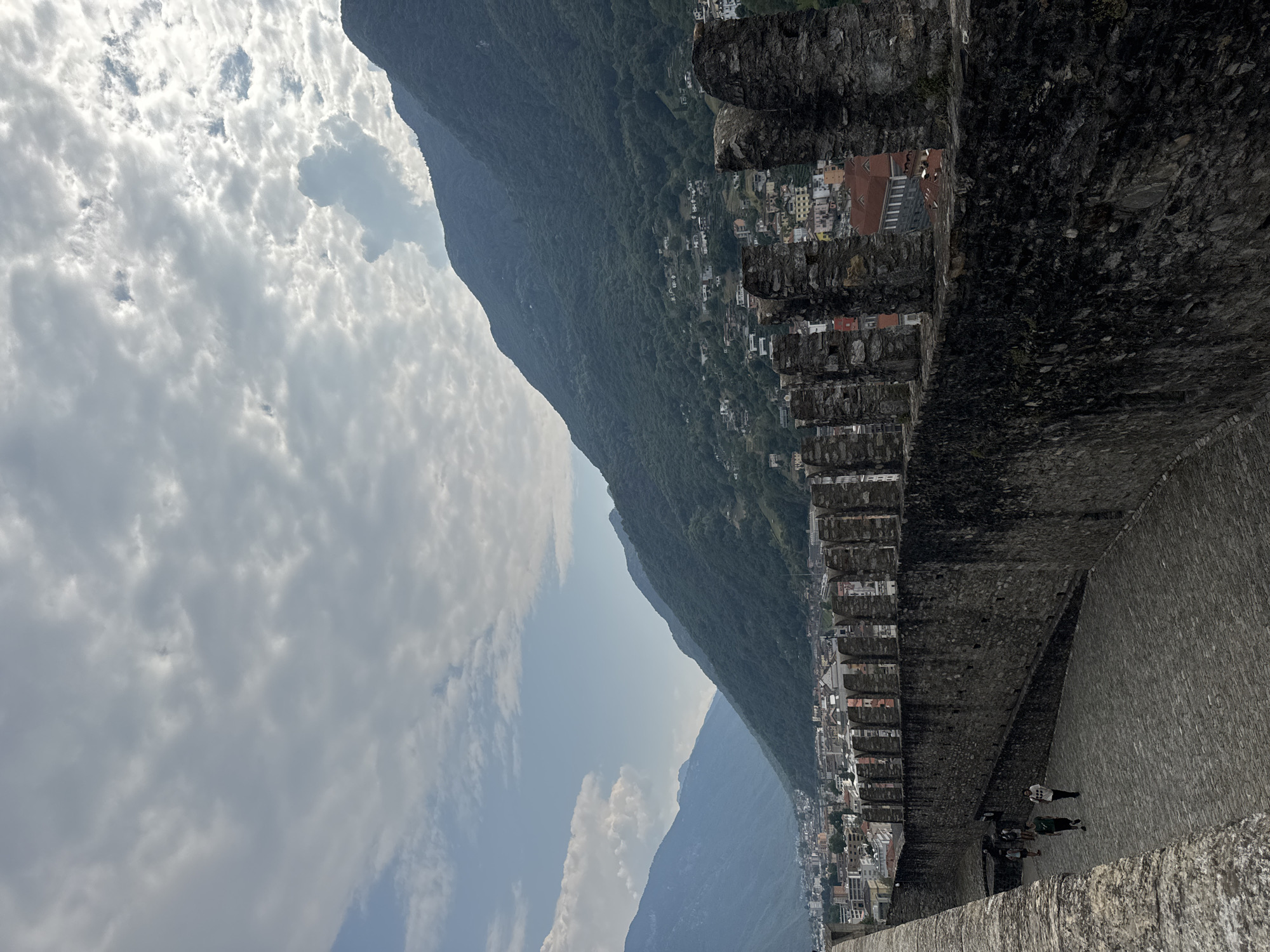 The image shows a view from the ramparts of Castel Grande in Bellinzona, Switzerland. The stone walls of the castle are in the foreground, with crenellations lining the top. A cobblestone pathway runs along the ramparts, where four individuals are walking away from the camera. In the midground, a town with buildings of various colors and architectural styles is nestled between two lush green mountains. The sky above is filled with dramatic, textured clouds. The overall impression is one of historical grandeur and natural beauty.
