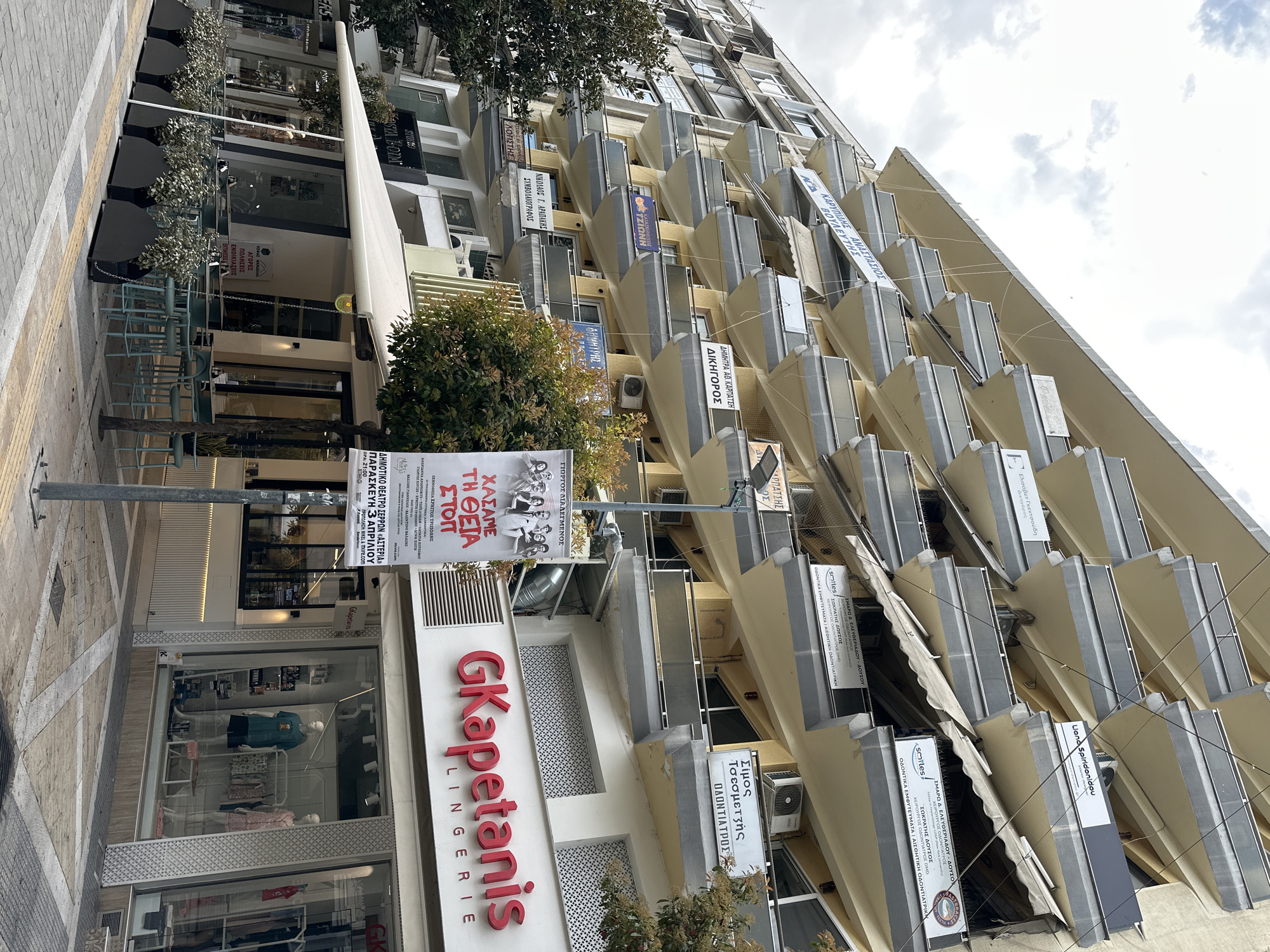 The image shows a street scene in Serres, Greece, featuring a multi-story building with a distinctive facade of protruding, angled architectural elements. These are adorned with numerous signs, mostly in Greek, displaying various business names and professions, such as lawyers, dentists, and real estate agents.
In the foreground, on the left, is a sidewalk with trees and a pole supporting a poster for a theatrical production. To the right is the front of a shop, Gkapetanis Lingerie, with mannequins displayed in the window. The shop is identified by the large red lettering spelling out “GKapetanis Lingerie” above its entrance.
The sky is overcast, suggesting a cloudy day. The perspective is from a street-level position, angled upwards to capture the height of the building. The overall impression is of a bustling commercial street.