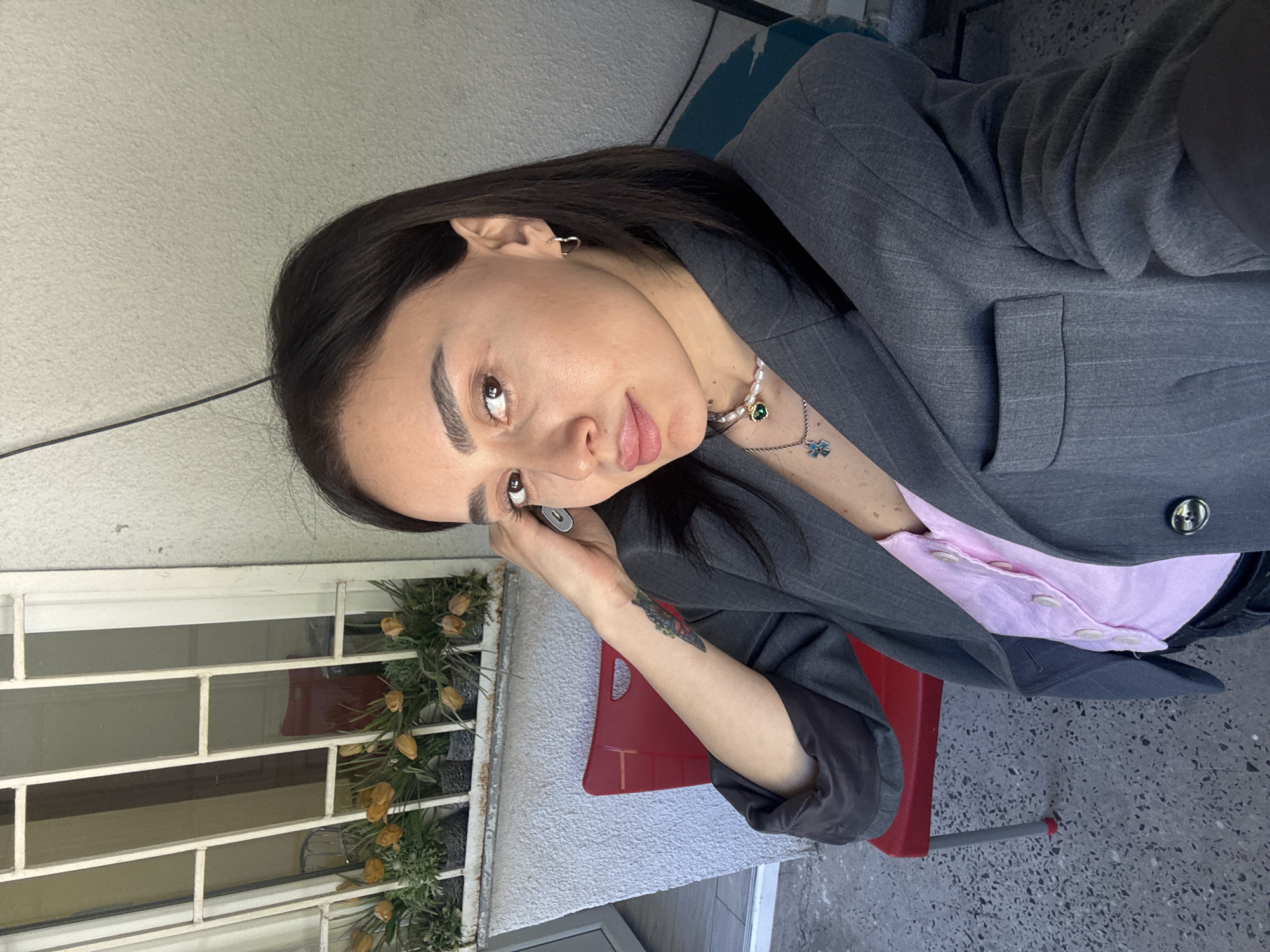 A woman with dark hair, wearing a gray suit jacket over a light pink shirt and a pearl necklace, sits on a red chair outdoors. She has a tattoo on her left forearm and is holding a phone to her left ear. A window with decorative plants is behind her to the left. The location appears to be Tbilisi, Georgia, based on the architecture and the general urban landscape. The ground is a speckled gray surface.