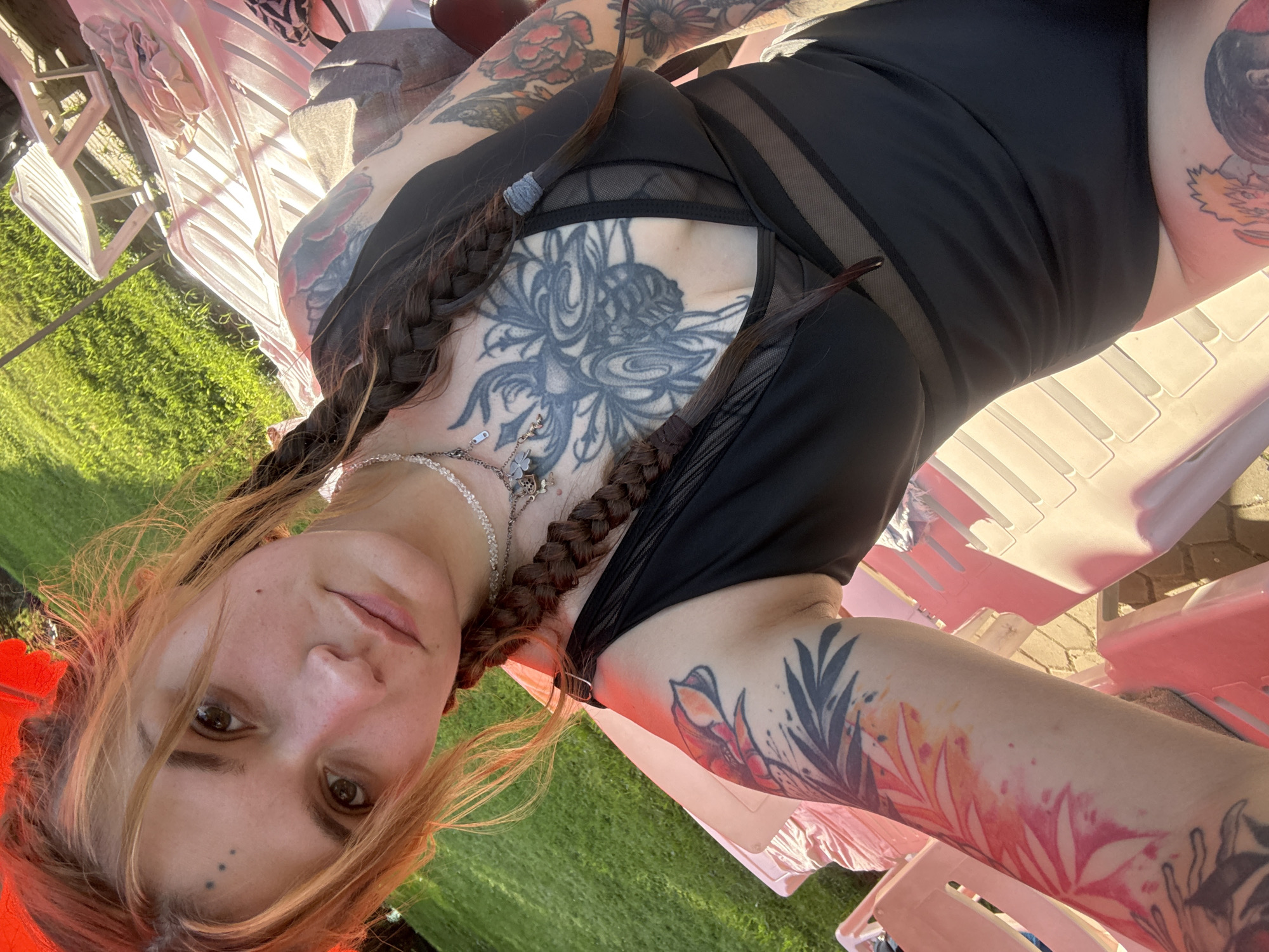 A woman with reddish-blonde hair, styled in two braids, is pictured outdoors. She is wearing a black swimsuit with mesh detailing on the chest. She has extensive tattoos covering her chest, arms, and visible on her leg. One tattoo on her chest features a large, intricate design, while her arms are adorned with floral and botanical motifs in vibrant colors, as well as some black ink designs. She is wearing a silver necklace. Her face is tilted upwards, and she has light brown eyes and fair skin, with a few small beauty marks on her face.
The setting appears to be a backyard or patio area, with green grass and what looks like a paved walkway visible. There are pink plastic chairs in the background, some of which are casting shadows, suggesting it is daytime. The lighting indicates it is likely late afternoon or early evening due to the warm, directional sunlight. The overall impression is of a casual, relaxed outdoor environment. No text is visible in the image.
