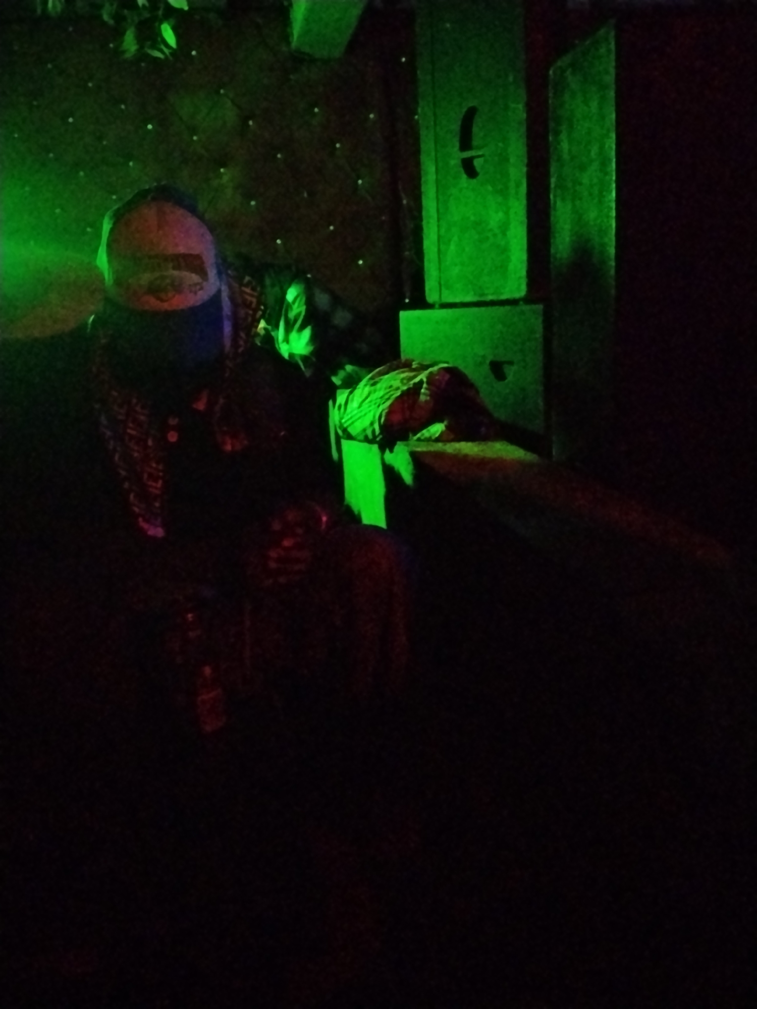 A person wearing a cap and a bandana over their face sits in a dark room illuminated by green light. The person is seated on a dark-colored surface, possibly a sofa. To the right of the person are two tall, green-lit rectangular objects stacked vertically, with cutouts on their front surfaces. The wall behind the person has small, scattered white lights. A patterned fabric is draped over the armrest of the seating, partially covering the person's right side. The overall scene is dimly lit, with significant portions of the image in shadow. The location is identified as Durimi, Nigeria.