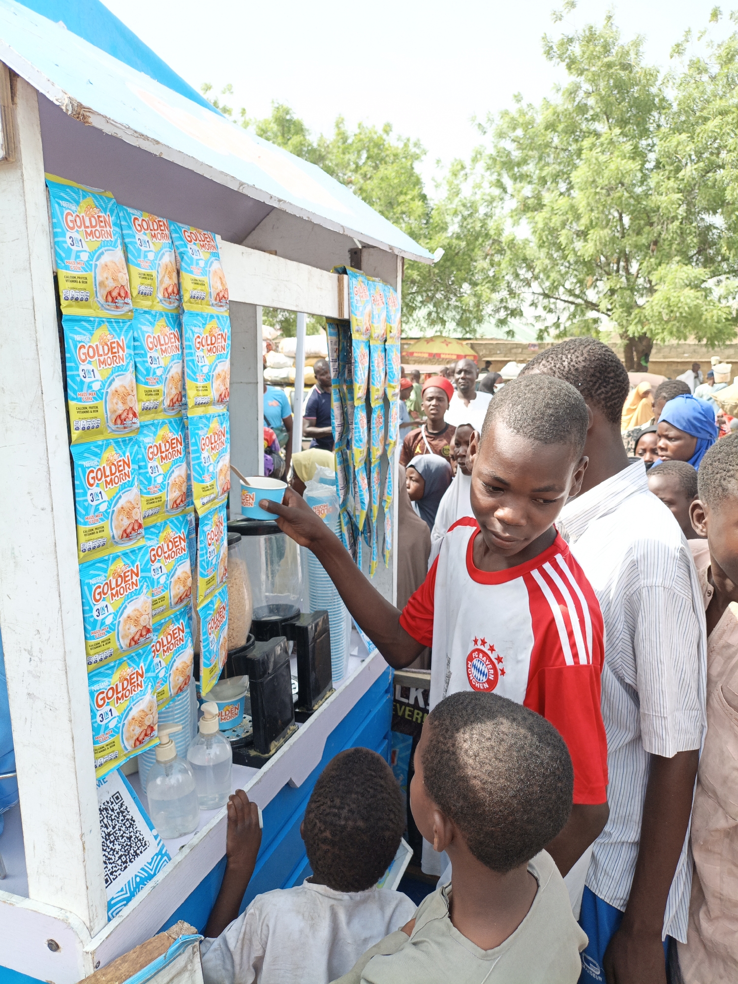 The image shows a vendor stand in Kawaji, Nigeria, selling Nestle Golden Morn 3 in 1 cereal in individual blue and yellow packages. Numerous packages are displayed. A line of people, mostly children, are gathered around the stand. A young boy in a white and red FC Bayern Munchen jersey is pointing towards the vendor stand. A clear plastic dispenser with a pump is visible, along with small plastic cups. The stand is painted white with blue accents, and a QR code is visible on its side. The environment includes trees and other people in the background.