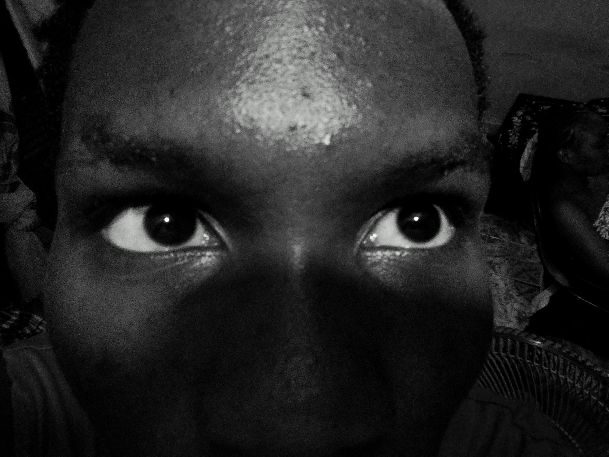 A black and white close-up image focuses on a person's face, primarily the eyes, forehead, and nose. The person has dark skin, with visible pores on the forehead. The eyes are wide open, reflecting light. The background is indistinct, with blurred objects including what appears to be a fan. A person is partially visible to the right side of the scene. Location: Unknown, Unknown.
