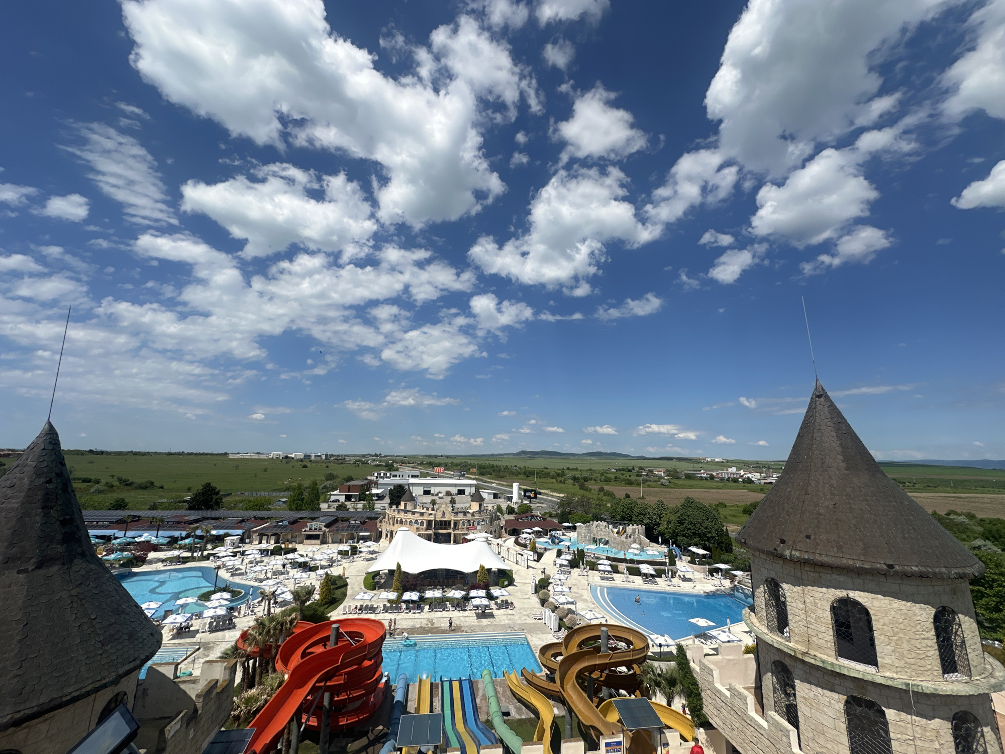 The image shows a view from atop a building at the Aquapark Nessebar water park in Ravda, Bulgaria on a sunny day with puffy white clouds in a bright blue sky. The view is of the waterpark with various pools, waterslides, buildings, lounge chairs, and umbrellas. In the distance is a green field, buildings, and roads. Two castle-like structures frame the scene in the lower left and right corners. The waterpark itself has a large white tent-like structure. People are seen in the pools and walking around the park.