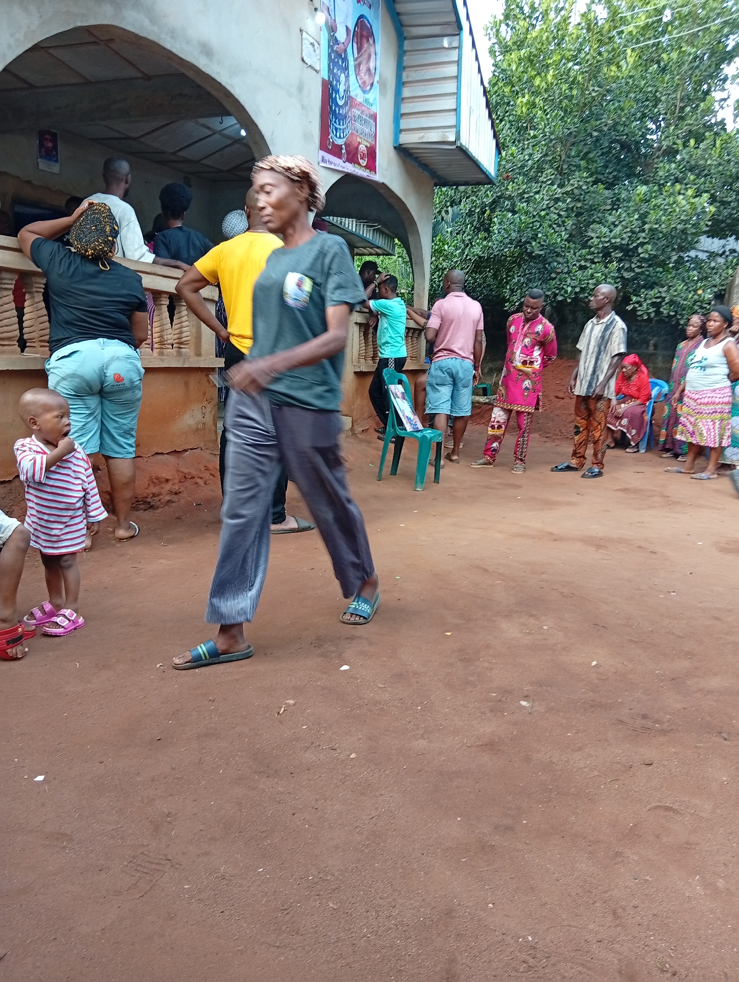 The image captures a scene in what appears to be a residential area. A group of people are gathered in front of a building with an arched veranda. The ground is bare earth. A woman walks towards the viewer. A child in a striped shirt is visible on the left. The exact location is unknown, as the city is specified as Unknown, Unknown.
