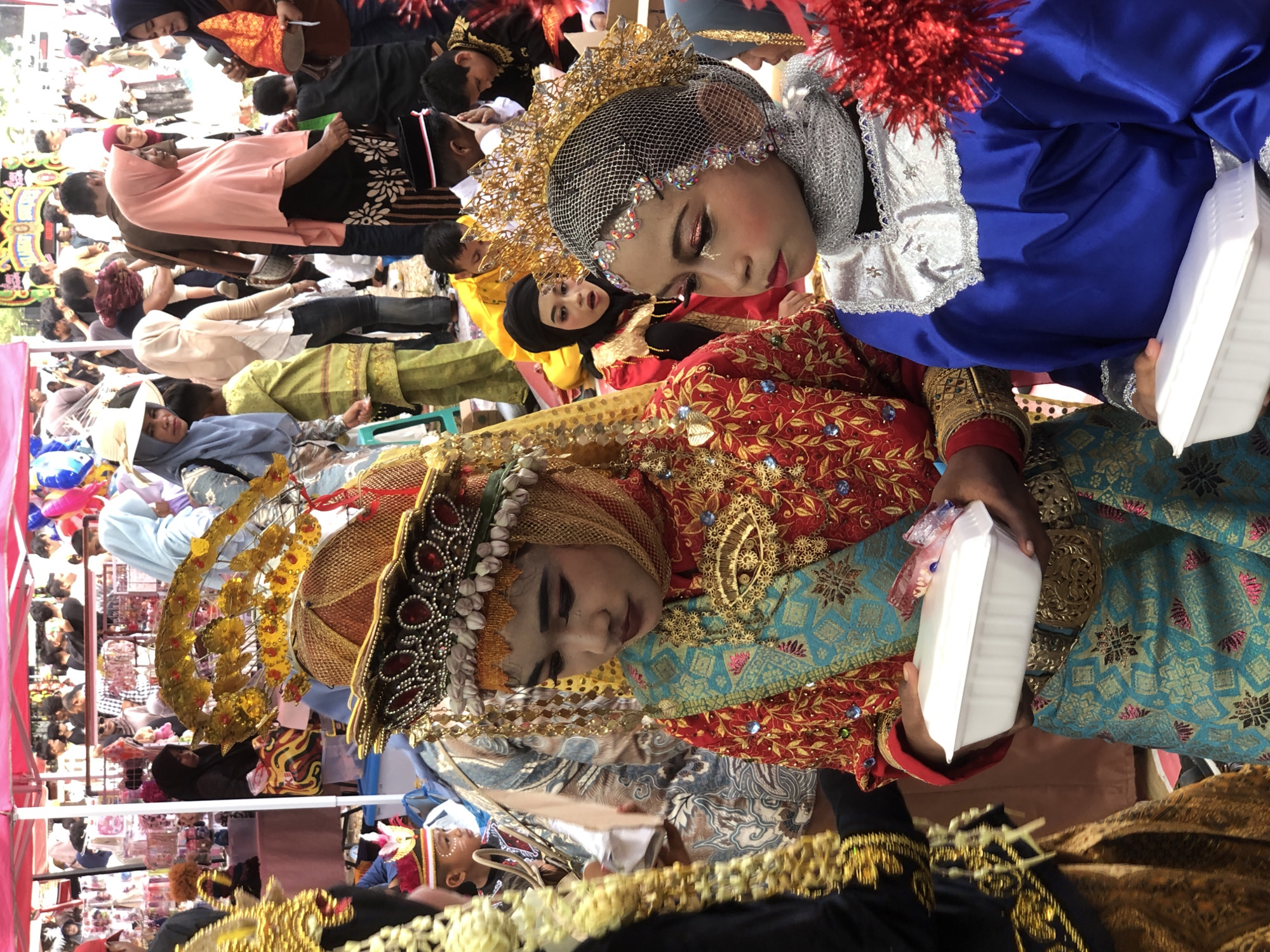 In Lhokseumawe, Indonesia, this outdoor event captures a vibrant procession or gathering. In the foreground, two young girls are adorned in elaborate traditional attire. The girl on the left wears a red and gold embroidered top with a turquoise patterned skirt and an ornate gold headdress. She holds a white rectangular container. The girl on the right is dressed in a bright blue robe with silver trim and a matching silver head covering. She also holds a white rectangular container. Both girls have heavily applied makeup. Behind them, other individuals in traditional dress and casual clothing are visible. The background features a red tent structure and stalls selling various items, suggesting a marketplace or festival setting. The scene is bustling with activity, indicating a community event in the city.