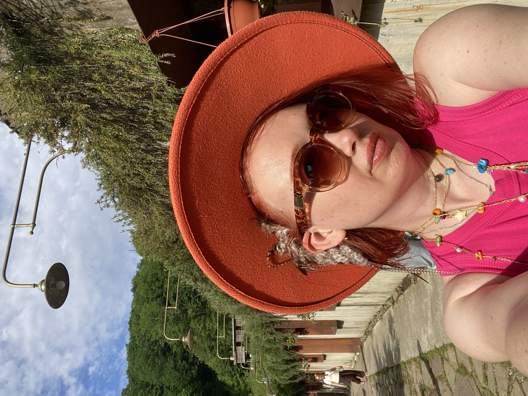 A woman wearing an orange hat, sunglasses, a pink top, and necklaces is posing outdoors. She has reddish-brown hair and fair skin. Her left arm is visible in the lower right corner. She is wearing feathers around her hat. In the background, there are trees, street lights, and a concrete walkway with wooden dividers. There is a person sitting on a bench in the distance. The sky is blue with white clouds. The location of the image is not evident.