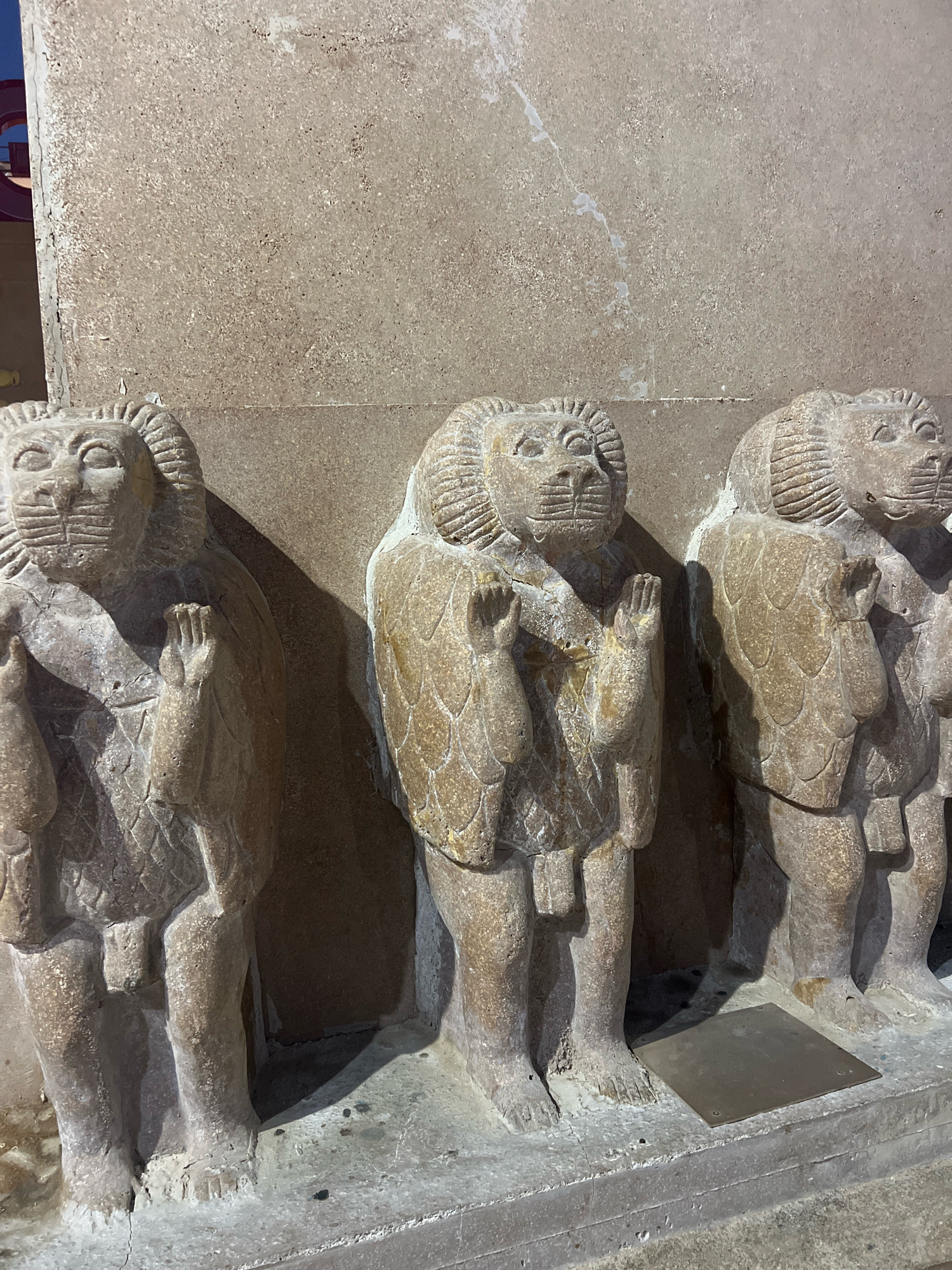 The image shows three ancient stone statues of lion-headed humanoids standing in a row. The statues are carved from a light-colored stone and appear to be weathered. Each statue has a lion's head with a mane, human-like arms raised in a gesture of prayer or supplication, and a human torso. They are wearing what appear to be patterned garments. The statues are positioned in front of a rough stone wall. The scene is illuminated by artificial lighting, casting shadows. There are no people visible in the image. This scene is located in the city of Sharm El Sheikh, Egypt.