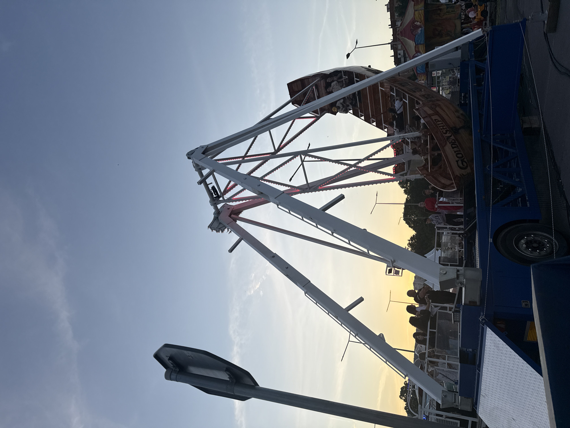 The image depicts a lively fairground scene in Shumen, Bulgaria, during the golden hour or dusk. A prominent "pirate ship" style swing ride, called "GOLDEN SHIP," is the central feature. The ride's boat-shaped carriage, made of what appears to be wood, is filled with numerous people who are either riding or waiting to ride. The robust white metal support structure of the swing ride is illuminated with rows of red lights, suggesting an evening event.
The sky transitions from a clear, light blue overhead to a warm, soft orange and yellow hue closer to the horizon, indicative of the setting sun. Some faint, wispy clouds are visible in the upper sky. Around the base of the ride, several individuals can be seen standing, potentially observing or waiting for their turn. The ride's blue base structure, equipped with large wheels, confirms its mobile fairground nature. In the background, hints of other carnival attractions, tents, and lighting fixtures are visible, contributing to the festive atmosphere. A streetlight appears in the foreground and another further back, illuminating parts of the scene. The overall impression is one of leisure and entertainment at an outdoor event.