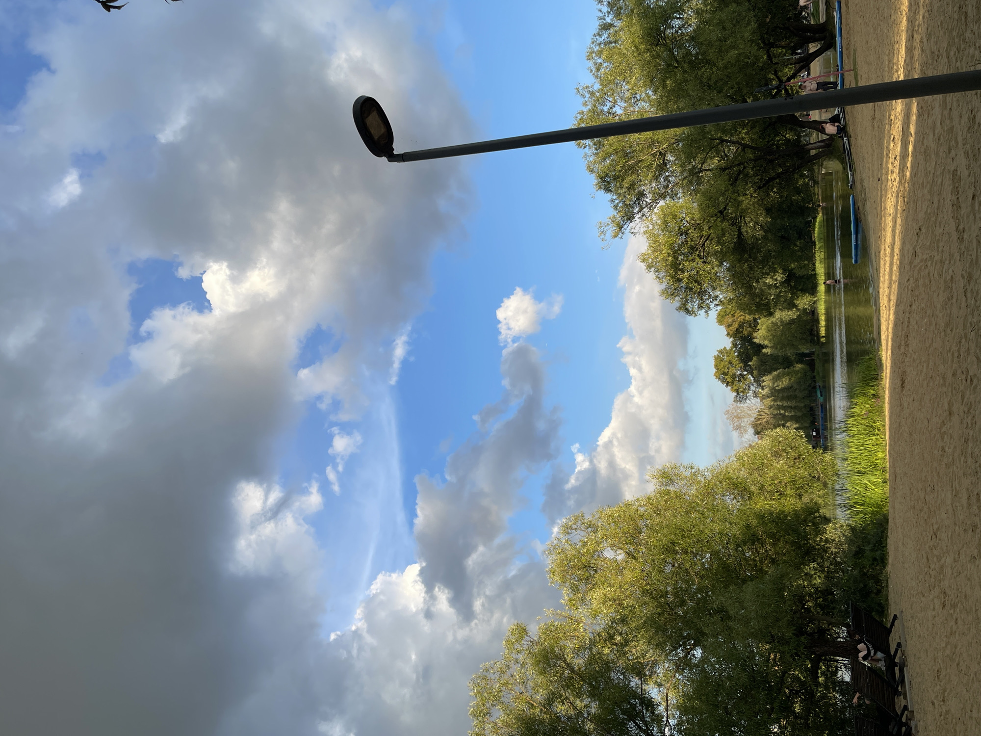 The image depicts a sandy beach beside a body of water, likely a lake, with lush green trees bordering the shore. The sky is a mix of bright blue and dramatic grey clouds, suggesting either a transition in weather or early evening lighting. In the foreground, there is a dark lamppost to the right of the frame, and a few lounge chairs on the sand to the left. Several people are visible in the water, some appearing to be swimming or paddleboarding, while others are relaxing on the shore. The overall atmosphere suggests a recreational area within a park or natural setting. The specific location is in the city of Unknown, Unknown.