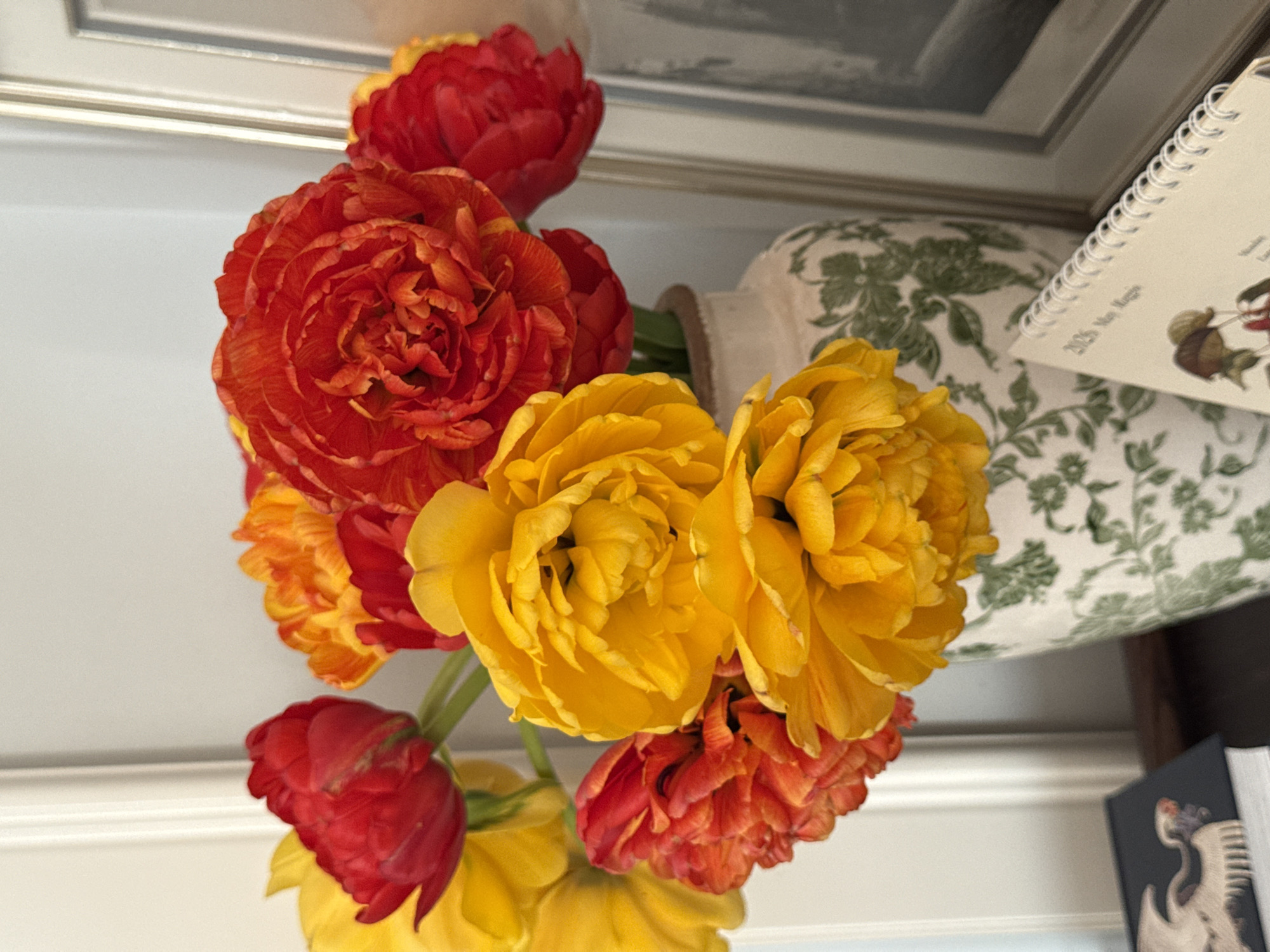 The image shows a bouquet of red, orange, and yellow flowers in a vase. A calendar and framed picture sit in the background. The setting is a room, possibly in a private residence. The location cannot be identified as Moscow, Russia, based on the visual information.