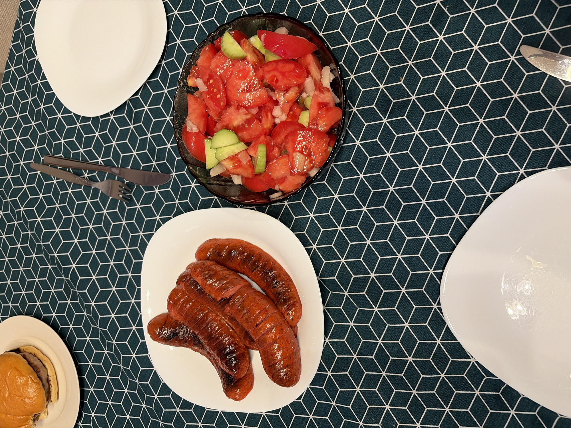 A dining table in Tsarevo, Bulgaria, is covered with a dark green tablecloth patterned with interlocking white geometric cubes. On the left, a white, irregularly shaped plate contains five grilled sausages exhibiting charring. Above and to the left of the sausages, another white plate holds a hamburger with a golden-brown bun, partially visible. To the right of the sausages, a clear glass bowl is filled with a fresh salad of chopped red tomatoes, sliced green cucumbers, and diced white onions, with hints of red bell pepper. An empty white, irregularly shaped plate is positioned on the upper right, accompanied by a silver fork and knife laid parallel. Another empty white, irregularly shaped plate is visible at the bottom, with a partial view of a silver knife to its lower right. The scene is indoors and well-lit, prepared for a meal.