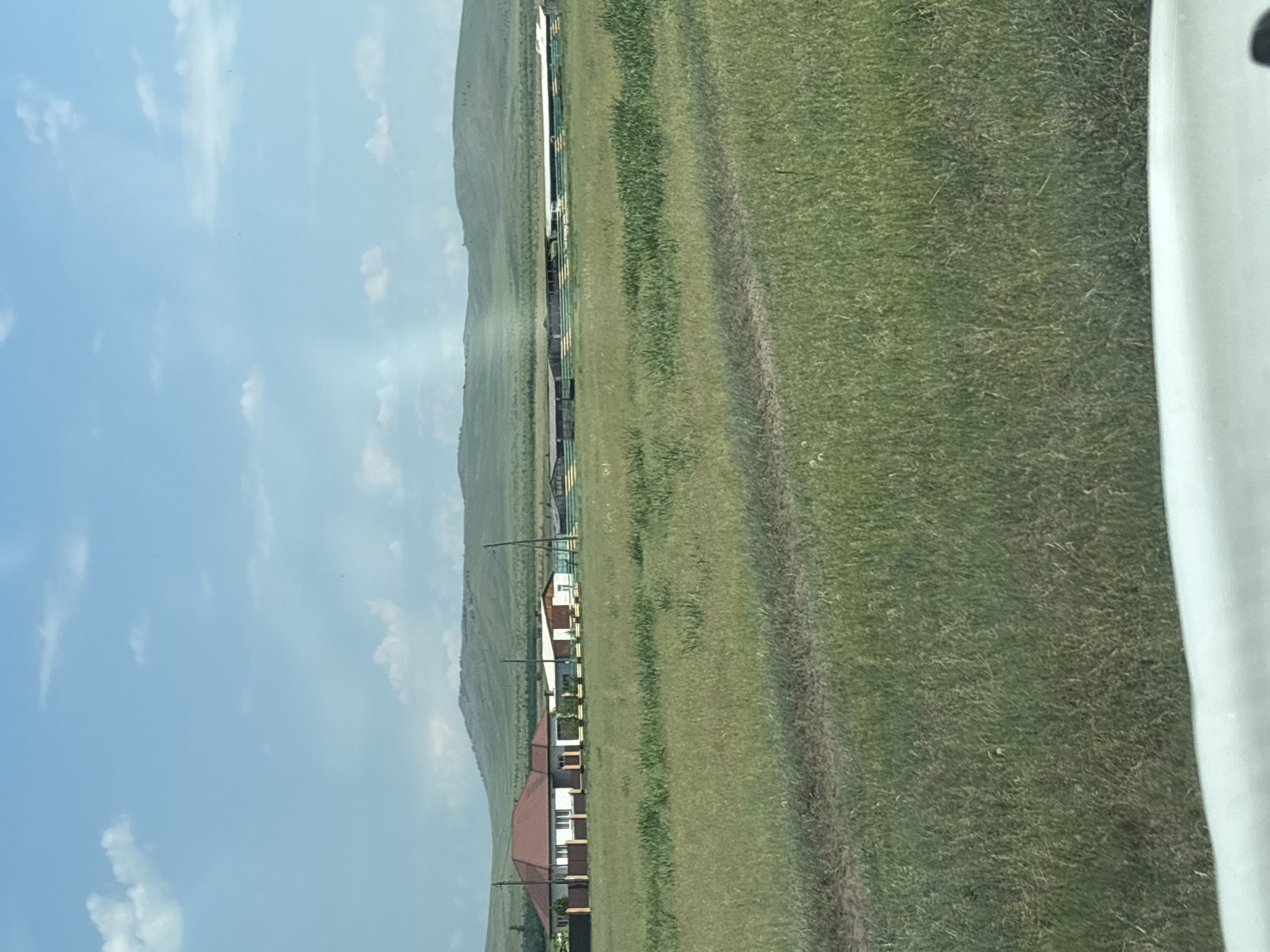 The image depicts a rural landscape in the city of Troitskoye, Russia. The scene features a long, low-lying building with a green and yellow striped facade, possibly a stable or agricultural structure, situated in the middle ground. To the left, there is a residential house with a reddish-brown roof and white walls, accompanied by a fenced enclosure. The foreground is dominated by a vast expanse of green grassland with a dirt track running through it. In the background, rolling hills under a partly cloudy blue sky complete the vista. No people or animals are visible in the frame.