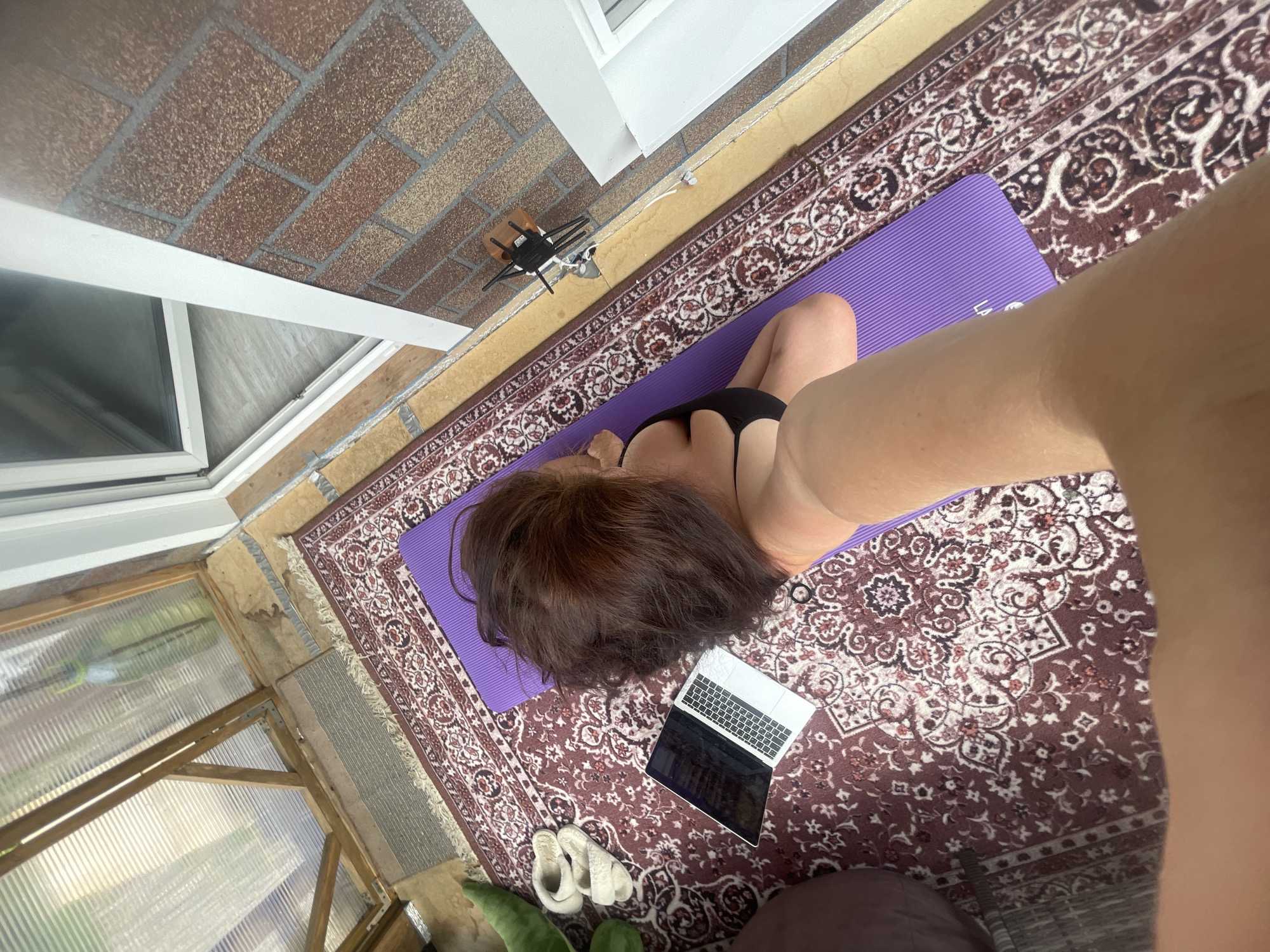 A high-angle shot shows a woman in a black bikini top and a purple yoga mat on a patterned rug. A laptop is open on the rug near her head, and a pair of white slippers are nearby. The location appears to be a sunroom or enclosed porch with brick walls and a window. In the top left corner, there is a window with a white frame and brick wall. In the bottom left corner, there is a window with a wooden frame and green outdoor scenery outside. A small black chair is placed against the brick wall. The location may be in the city of Мосметаллоконструкция Онт, Russia.