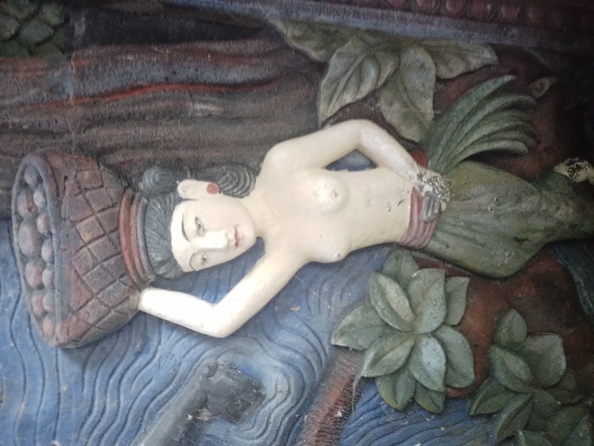 The image depicts a bas-relief sculpture of a woman lying on her side, possibly in water, with a basket or container beside her head. She is topless and has a decorative waistband. Foliage surrounds her. The relief is likely part of a larger structure or mural, but its specific context or story is unknown. This bas-relief sculpture is located in Kuta Selatan, Indonesia.