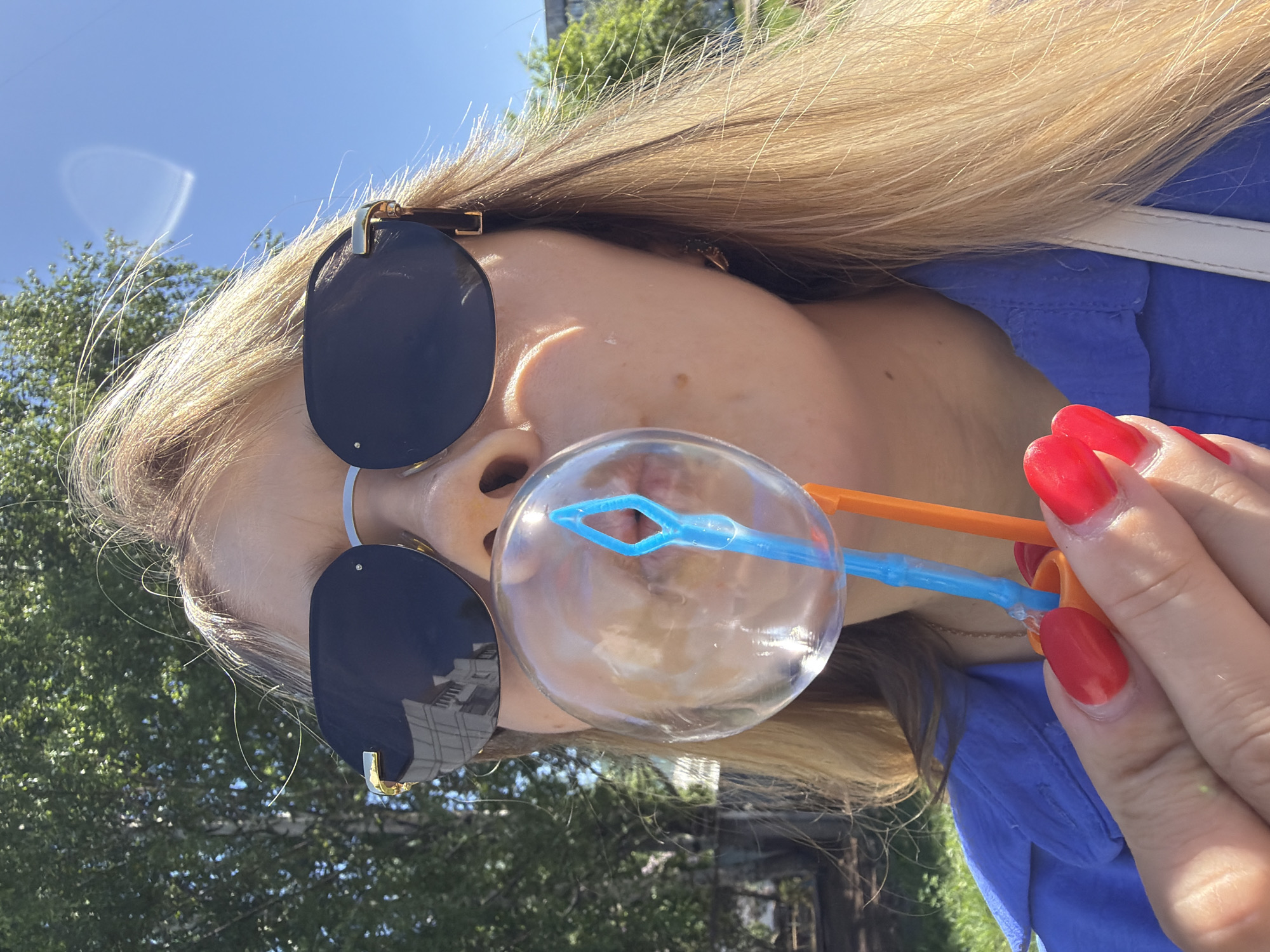 A woman is blowing a bubble in an outdoor setting. She is wearing sunglasses, has blonde hair, and her nails are painted bright red. She is holding a blue and orange bubble wand. She is wearing a blue shirt. The background shows green trees and a building reflecting in the sunglasses. Based on the metadata of the image, this photograph was taken in Chaikovsky, Russia.