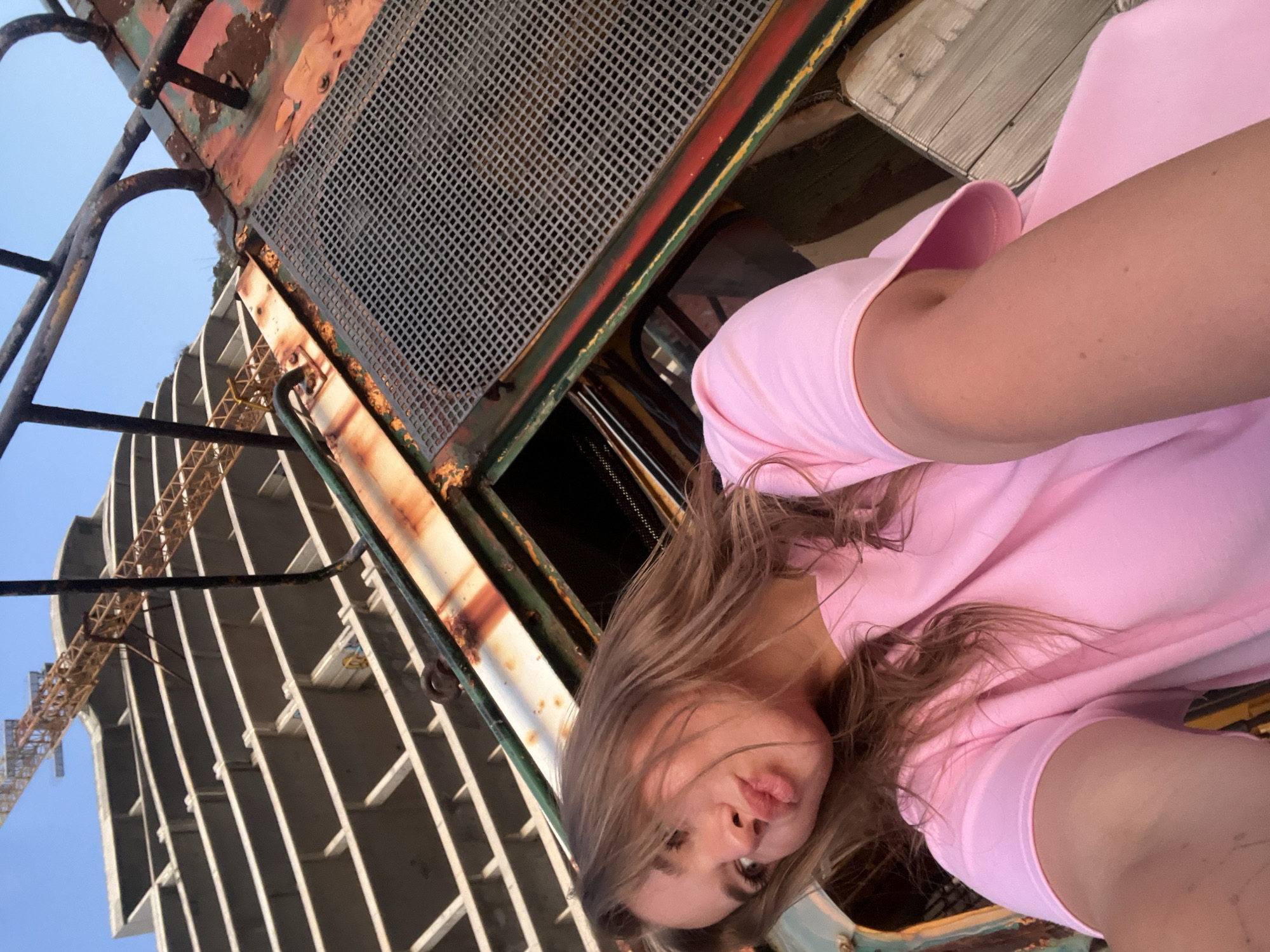 A photograph shows a woman posing in front of a rusted train car with a building under construction visible in the background. The woman is wearing a pink shirt. The location is Perazića Do, Montenegro.