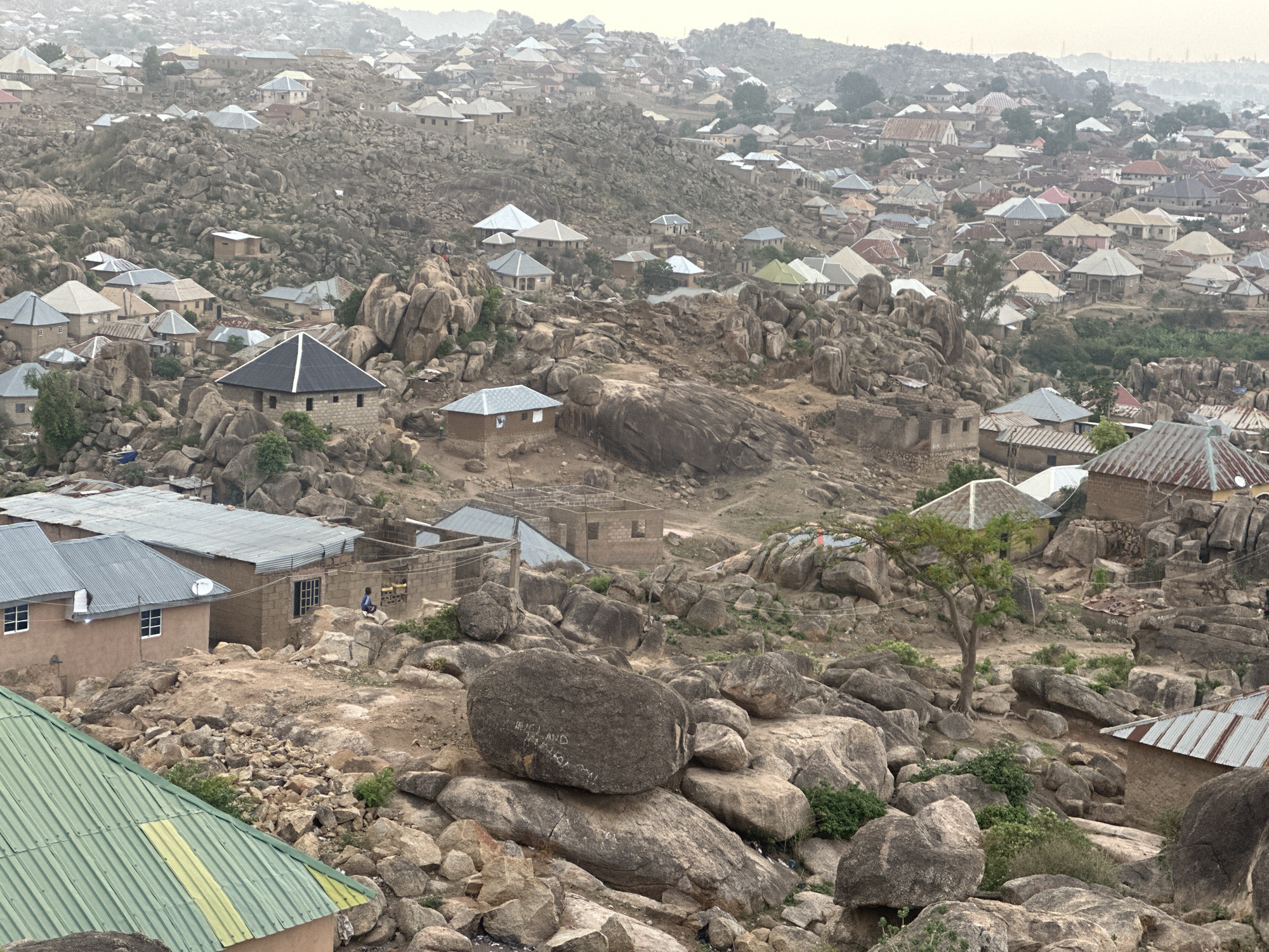 The image shows a densely populated residential area in Jos, Nigeria, built amidst a rocky landscape. The scene is dominated by numerous dwellings with a variety of roofing materials, ranging from corrugated metal to tile. These houses are interspersed with large boulders and rock formations. The terrain is rugged and uneven. In the lower portion of the image, there is a cluster of buildings with green, grey and silver roofs. Towards the middle, one can observe a single tree standing among the rocks. The overall atmosphere suggests a densely populated community adapting to a challenging geological environment. The sky is overcast. In the middle ground, some buildings are under construction.
