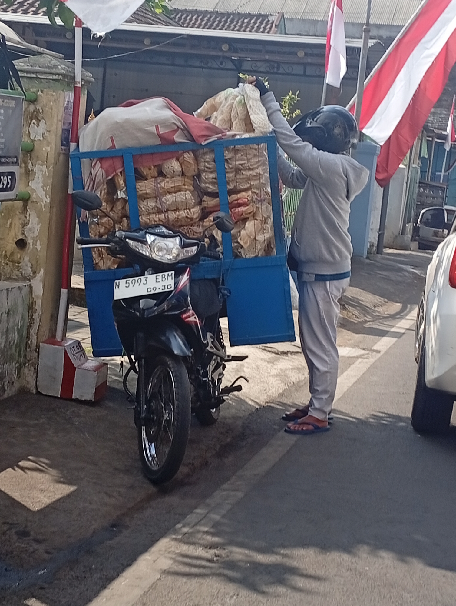 The image depicts a street scene in Malang, Indonesia, under bright daylight. A person wearing a grey hoodie, light-colored pants, a black helmet, and blue flip-flops is standing next to a motorcycle. The motorcycle, a black and silver model with a license plate reading "N 5993 EBM 09.30", is equipped with a blue metal cart filled with bagged goods, possibly food items, covered with a white and red cloth. The cart is attached to the back of the motorcycle.
In the background, there are buildings with tiled roofs and flags. One flag is prominently visible with red and white stripes, likely the Indonesian flag. To the right, the rear of a white car is partially visible. The ground is paved with asphalt and some concrete areas, with visible tire marks and shadows cast by the surroundings. A red and white striped marker post is located to the left of the motorcycle, adding to the urban context of the location. The scene appears to be a typical street view in a city like Malang, where such mobile vendors are common.