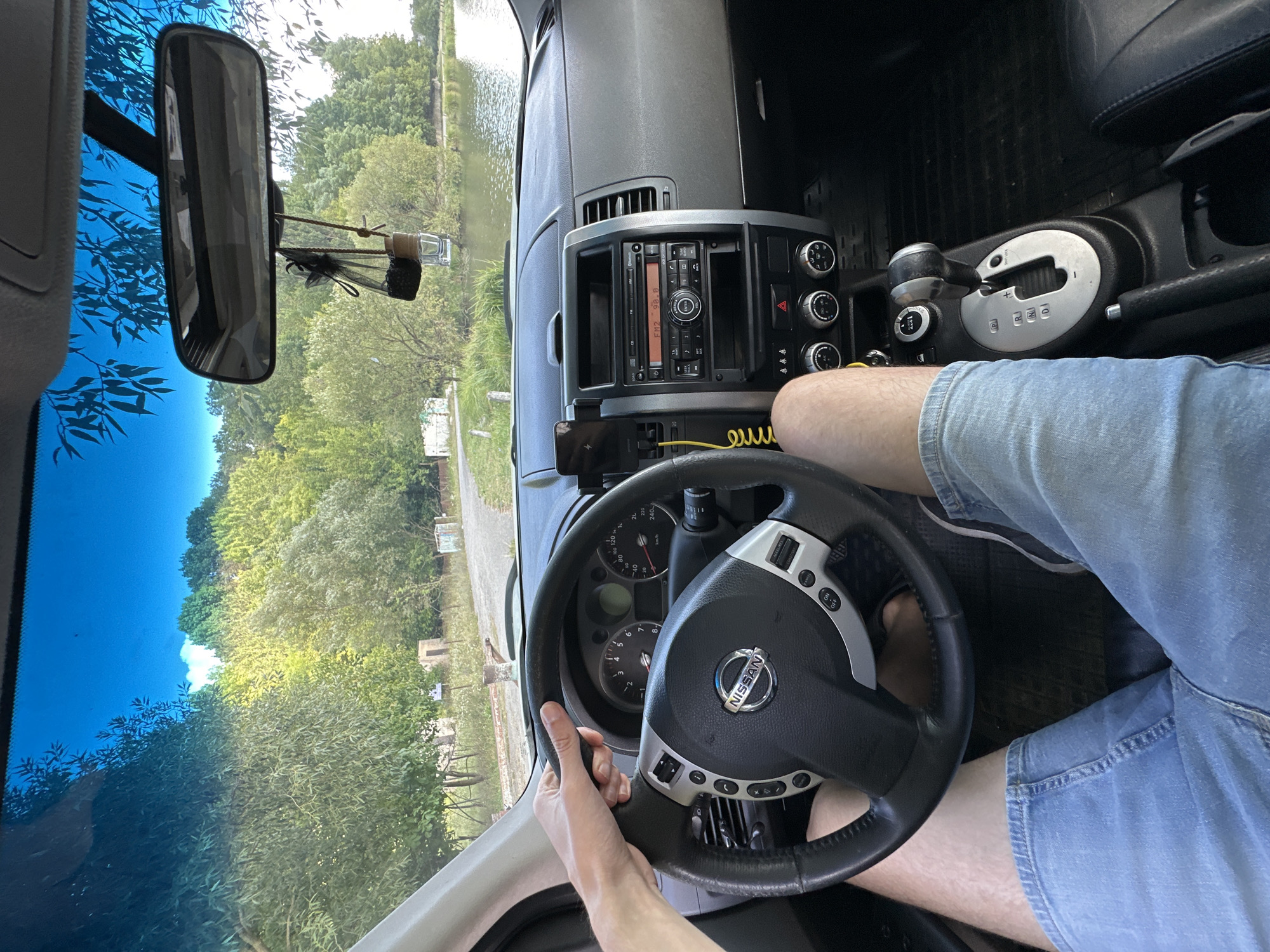 The image is taken from the interior of a car, looking out through the windshield and side windows. A person's hands are visible on the steering wheel of a Nissan, which is likely a Qashqai based on the dashboard design. The dashboard features a radio, climate controls, and gear shifter. The person is wearing blue denim shorts. Outside the car, a body of water, possibly a lake or river, is visible, with trees and greenery lining its banks. A road and some buildings are also in the background. The sky is blue with some reflections of tree branches on the windshield. The context of Makiv, Ukraine, suggests this is a rural or semi-rural setting. The image captures a moment of driving in a scenic, natural environment.