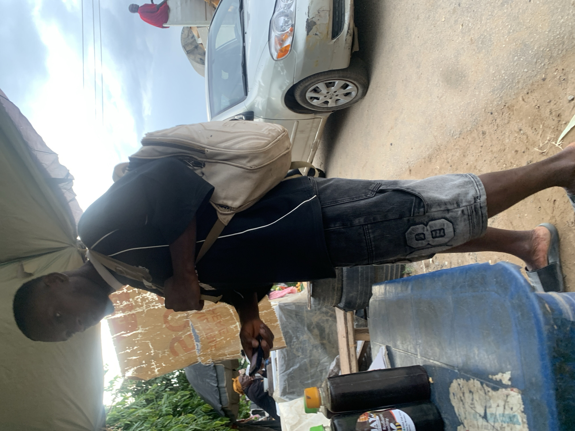 The image depicts an outdoor scene in Abuja, Nigeria, likely a market or roadside area. In the foreground, a man wearing a black t-shirt, denim shorts with a decorative patch, and sandals stands facing to the left, holding a mobile phone. He is also wearing a beige backpack. To his left, on a blue plastic table, are several bottles, one of which appears to be dark liquid with a label that reads "MAY". The table also shows signs of wear and tear. Behind the man, a silver sedan is parked on a dirt road. Further in the background, another person in a red shirt is visible sitting on the back of a truck, and a person wearing a yellow headscarf is seated under a black umbrella near some umbrellas. The environment appears somewhat dusty, with vegetation visible on the left side of the image and clouds in the sky.