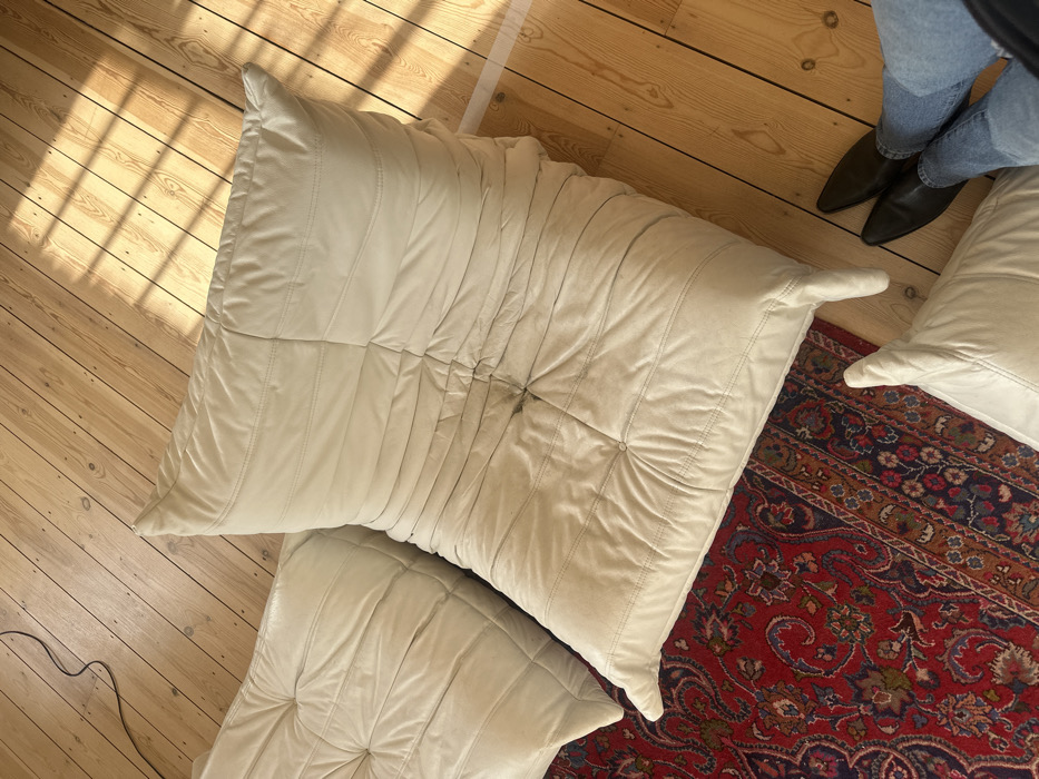 An interior scene depicts a living space within an unidentified residential or commercial property in London, United Kingdom. The foreground features three large, cream-colored modular sofa or floor cushions, consistent with a Togo design, distributed across a section of a red and blue patterned Oriental rug and light-toned wooden floorboards. The prominent central cushion displays visible stitching, characteristic pleating, and a localized dark stain or area of discoloration on its upper surface. Natural light from an unseen window projects distinct grid-patterned shadows onto the wooden floorboards on the right side of the frame. In the lower right, the lower legs of an individual are visible, clad in light blue denim jeans and black, pointed-toe ankle boots, positioned on the wooden floor near the cushions. A thin, dark electrical cord is present on the wooden floor in the upper left quadrant.