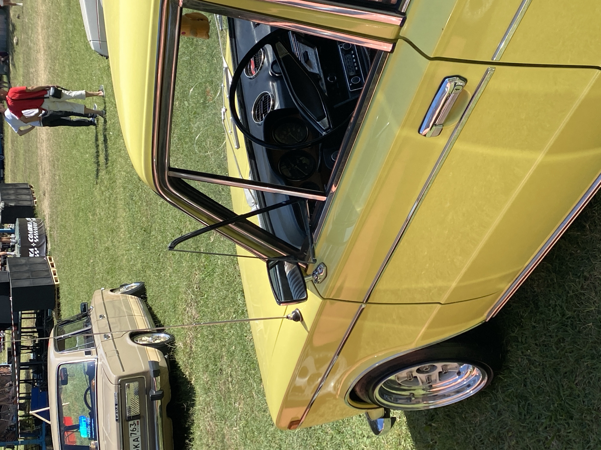 This image shows a yellow vintage sedan parked on a grassy field. The car is a classic Soviet-era automobile, likely a Lada. Its exterior features chrome accents and polished rims. The car's interior is partially visible, revealing a steering wheel and dashboard. In the background, several people are standing near a stage setup with large speakers, suggesting an outdoor event or festival. The environment appears to be a park or open field, with grass covering the ground. The overall scene depicts a relaxed, outdoor gathering with a focus on vintage automobiles. The location is Bezymyannoe, Russia.